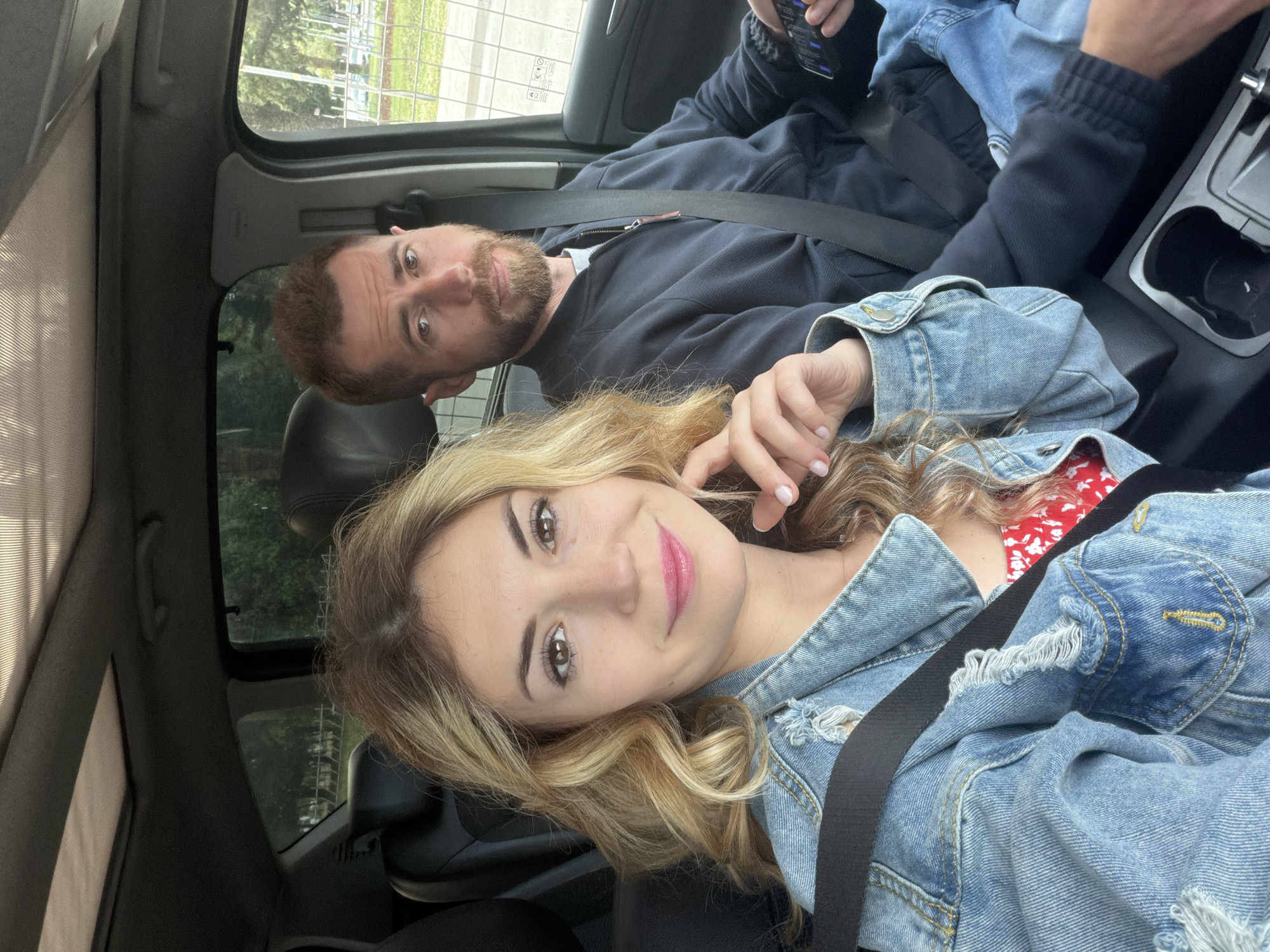 A man and woman are inside a car. The woman has blond hair and is wearing a denim jacket. She is in the foreground looking at the camera. The man has brown hair and a beard. He is in the background, looking at the camera. They are both wearing seatbelts. The car is parked, and the background shows trees and a fence, suggesting they are in an urban area but there is nothing to discern if they are in Budva, Montenegro.
