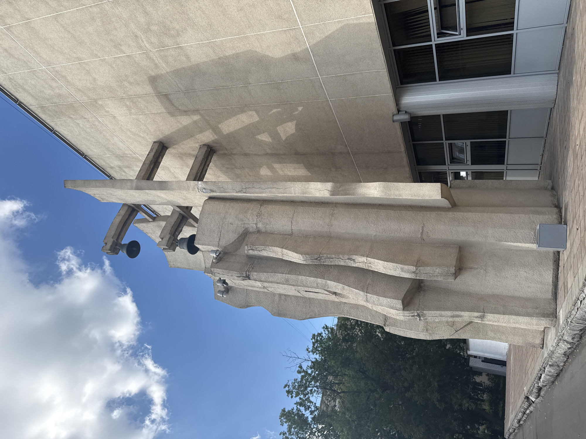 The image shows a concrete structure that appears to be part of a building's exterior or a sculpture. It is composed of large, angular concrete slabs with a textured surface, some of which are cracked. There are several concrete supports or beams extending from the structure. Attached to some of these supports are dark, bell-shaped objects, possibly speakers or bells.
Above the concrete structure, a large section of a building with a modern facade is visible. This section features a grid of windows and what looks like a corrugated metal awning or blind. The building's facade is made of glass and metal panels.
The scene is set against a bright blue sky with scattered white clouds. In the background, lush green trees are visible, and glimpses of other buildings can be seen.
The image is taken from a low angle, looking upwards. The lighting suggests it is daytime, with shadows cast by the concrete structure onto the building's overhang. The overall impression is one of an outdoor urban environment, possibly a public space or the exterior of a civic building. No people are visible in the image. There is no discernible text.