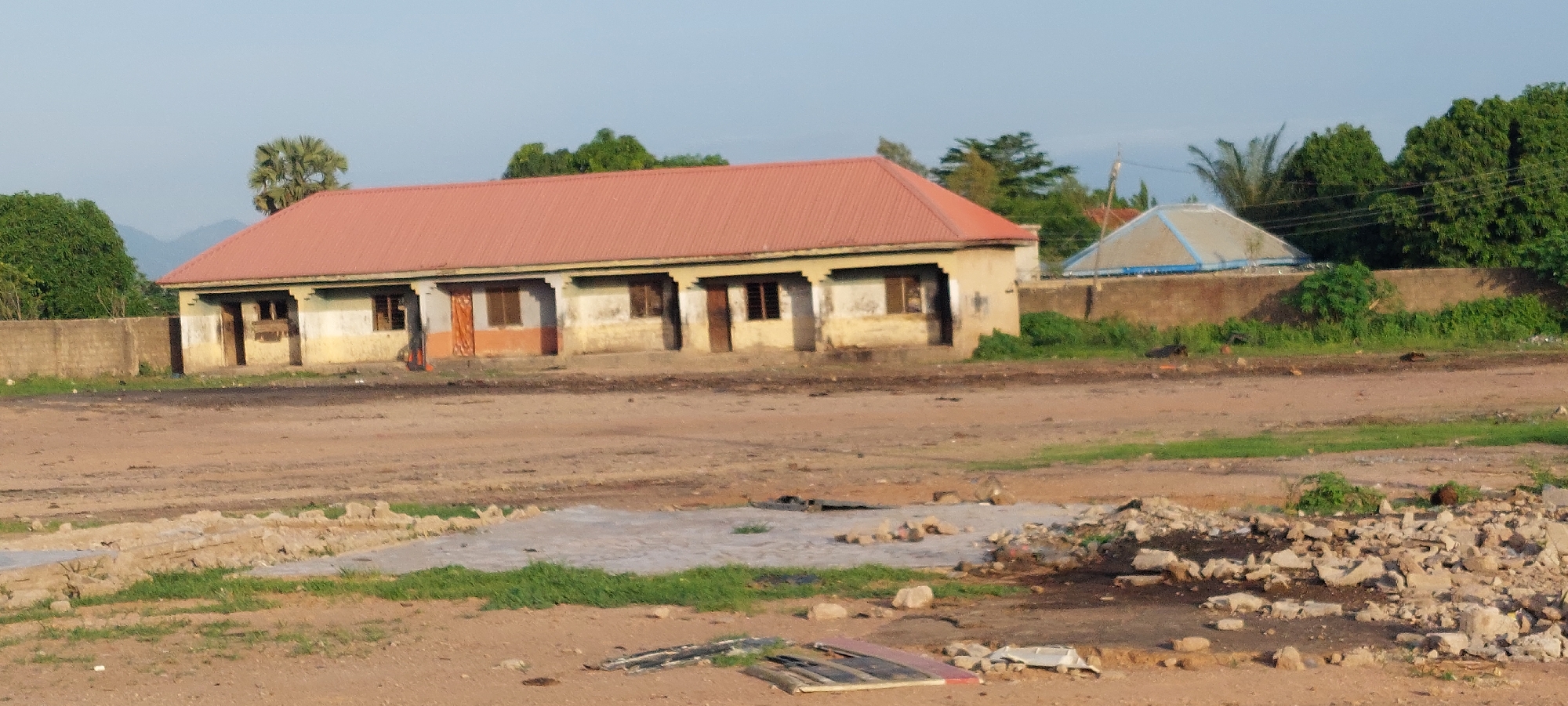 In Howayi, Nigeria, a long, single-story building with a red corrugated metal roof stands under a clear sky. The building, painted a pale yellow and white with some orange accents around doorways and windows, appears weathered. It has multiple doorways and windows, suggesting it may be a school, dormitory, or similar facility. In front of the building, a rough, dirt ground is visible. To the right of the main building, a geodesic dome structure is partially obscured by a low, brown wall and lush green vegetation. The surrounding environment includes more green trees, a palm tree, and a distant, hazy mountainous landscape. The foreground shows a rough, uneven terrain with scattered rocks and some concrete debris. The scene appears to be an outdoor, daytime setting.