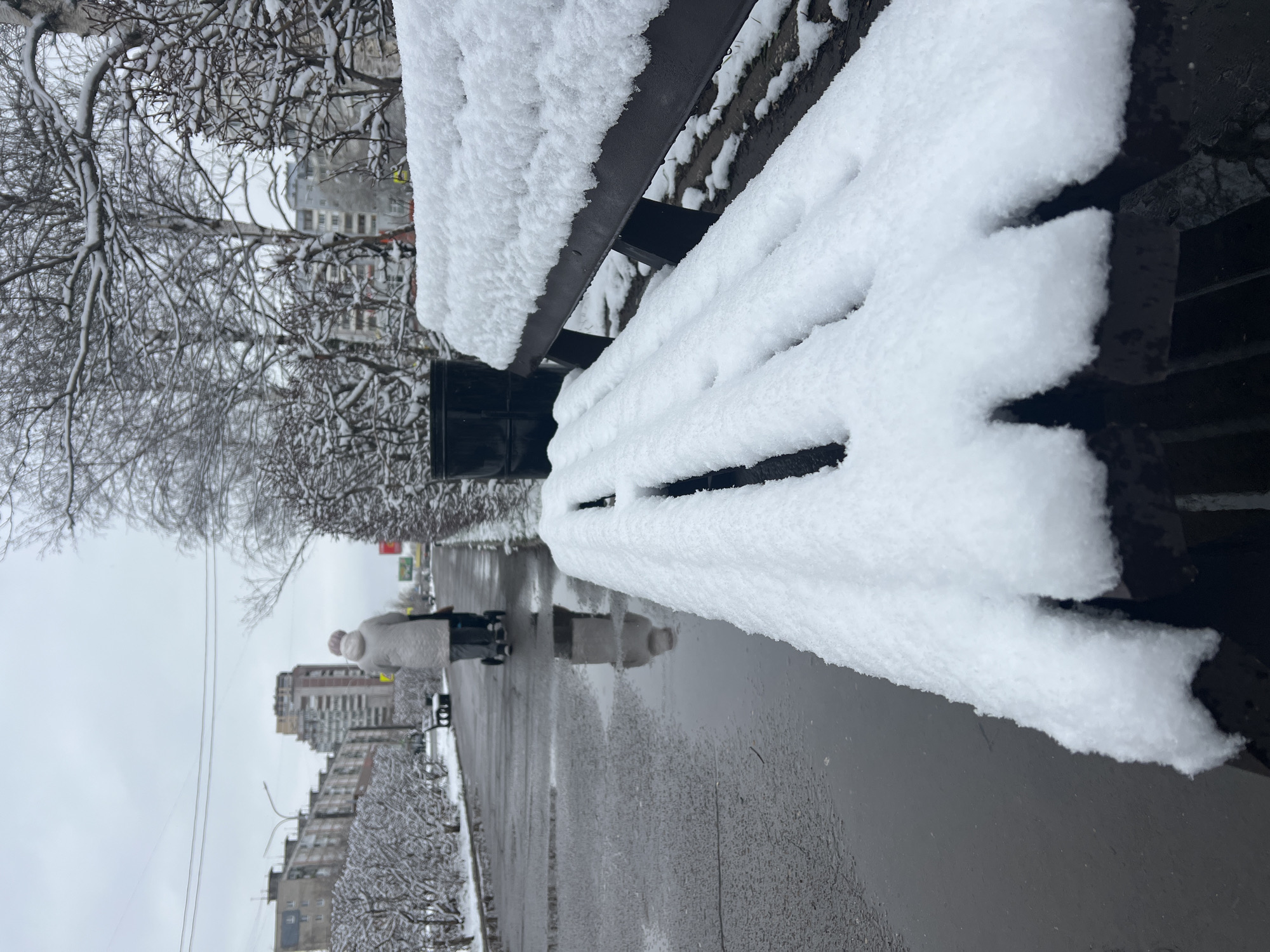 The image depicts a snowy scene in Perm, Russia. A snow-covered park bench dominates the foreground, with bare trees and buildings visible in the background. A person bundled in winter clothing walks along the sidewalk. The pavement is wet, reflecting the surrounding environment.