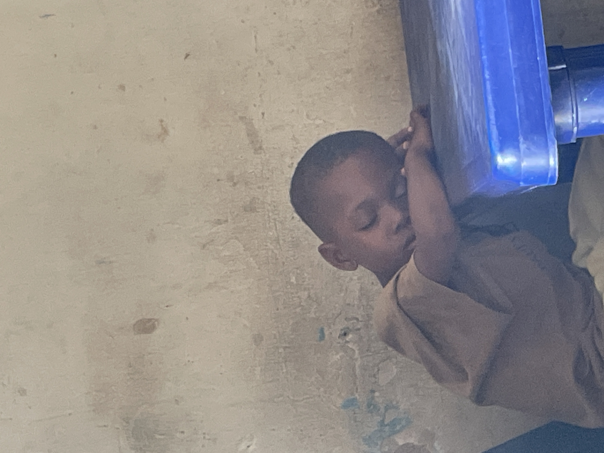 A young, dark-skinned boy is depicted holding a blue, rectangular object against his head. He has short hair and is wearing a light-brown or tan shirt. The background consists of a textured, light-colored wall. The location is Jos, Nigeria. The boy is physically supporting the blue object against the wall.