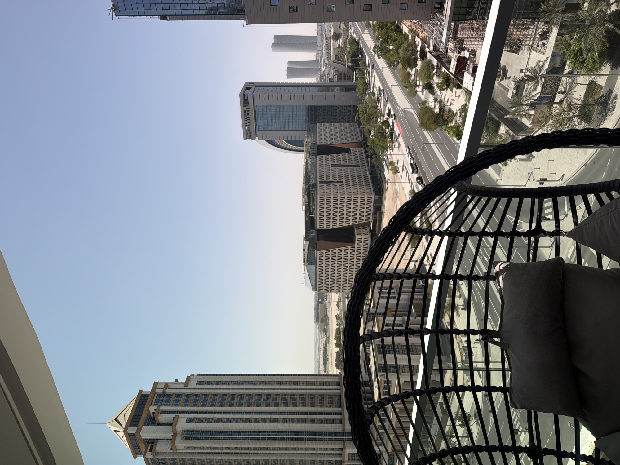 The image depicts a city skyline as seen from a balcony. In the foreground, a portion of a woven hanging chair with cushions is visible, suggesting a leisurely setting. The chair is dark in color and appears to be made of wicker or a similar material. Beyond the chair, the scene opens up to a wide view of a modern city.
Several tall buildings with glass facades and distinct architectural designs dominate the midground and background. Some buildings are rectangular, while others feature more complex geometric patterns. A wide road with multiple lanes runs through the city, with cars visible on it. Lined with trees, the road appears to be a major thoroughfare. The sky is clear and a pale blue, indicating daytime, likely mid-morning or afternoon due to the bright, diffused light. The overall atmosphere is one of urban development and modern architecture. There is no visible text in the image. The scene conveys a sense of calm and observation, as if someone is relaxing and enjoying the view from their balcony.