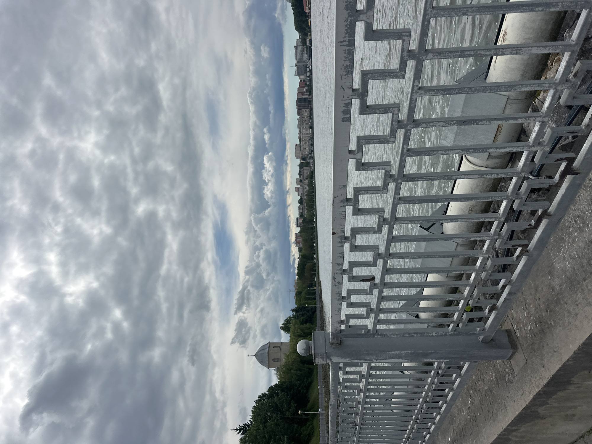 The image captures a panoramic view of Ternopil Lake in Ternopil, Ukraine, from an embankment under a heavily overcast sky. In the immediate foreground, a weathered grey metal fence with a repeating geometric pattern of interconnected squares runs horizontally across the frame. A prominent vertical fence post with a spherical finial is visible on the left side. Beneath the fence, a concrete walkway or embankment is present, and large, light-colored utility pipes, showing signs of wear and partial metal coverings, are visible along the water's edge, partially obscured by the fence bars.
The midground features the expansive, grey-toned surface of Ternopil Lake, exhibiting ripples indicative of wind or current. On the far bank, to the left, a dense line of green trees partially conceals the lower portions of structures. Above the treeline, a distinctive domed building with a pointed roof and a cross on top, identifiable as the Church of the Exaltation of the Holy Cross, stands prominently. Further along the far bank, a continuous urban skyline is visible, composed of numerous multi-story buildings, predominantly residential or commercial, stretching across the horizon. A construction crane is faintly discernible amidst the buildings on the far bank.
The background is dominated by a dense, grey cloud cover, indicating an overcast day. While most of the sky is obscured by varying shades of grey clouds, subtle patches of brighter sky and hints of blue are visible towards the horizon, particularly on the right side of the frame. No human figures or animals are present in the visible scene.