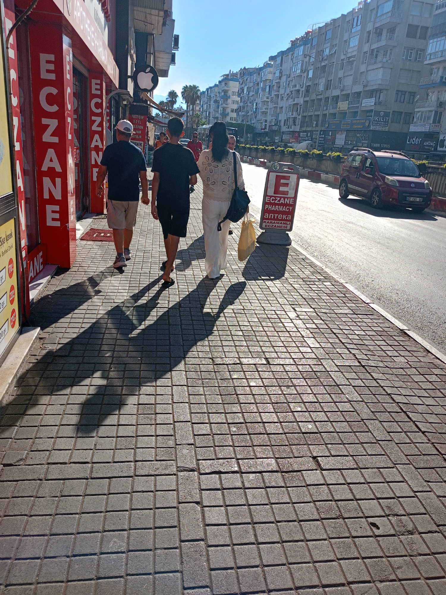 In Antalya, Turkey, three individuals walk away from the camera down a sun-drenched sidewalk paved with grey square tiles. The two men in front are wearing casual attire: one in a dark t-shirt and beige shorts, the other in a black t-shirt and black shorts, barefoot. The woman behind them wears a white, loosely fitting top and trousers and carries a light yellow shopping bag and a dark shoulder bag. They are walking past a row of shops. On the left, a store with bright red signage displays the word "ECZANE" vertically in white letters, repeated multiple times, indicating a pharmacy. Further down the street, a red sign with an "E" and the words "ECZANE PHARMACY" and "АПТЕКА" also marks a pharmacy. Beyond the sidewalk, a road runs parallel, with a red hatchback car parked and another vehicle driving away. The background is dominated by a long, multi-story building with numerous balconies, characteristic of urban architecture. Palm trees are visible further down the street, suggesting a Mediterranean climate. The bright sunlight casts long, distinct shadows of the pedestrians across the tiled sidewalk.