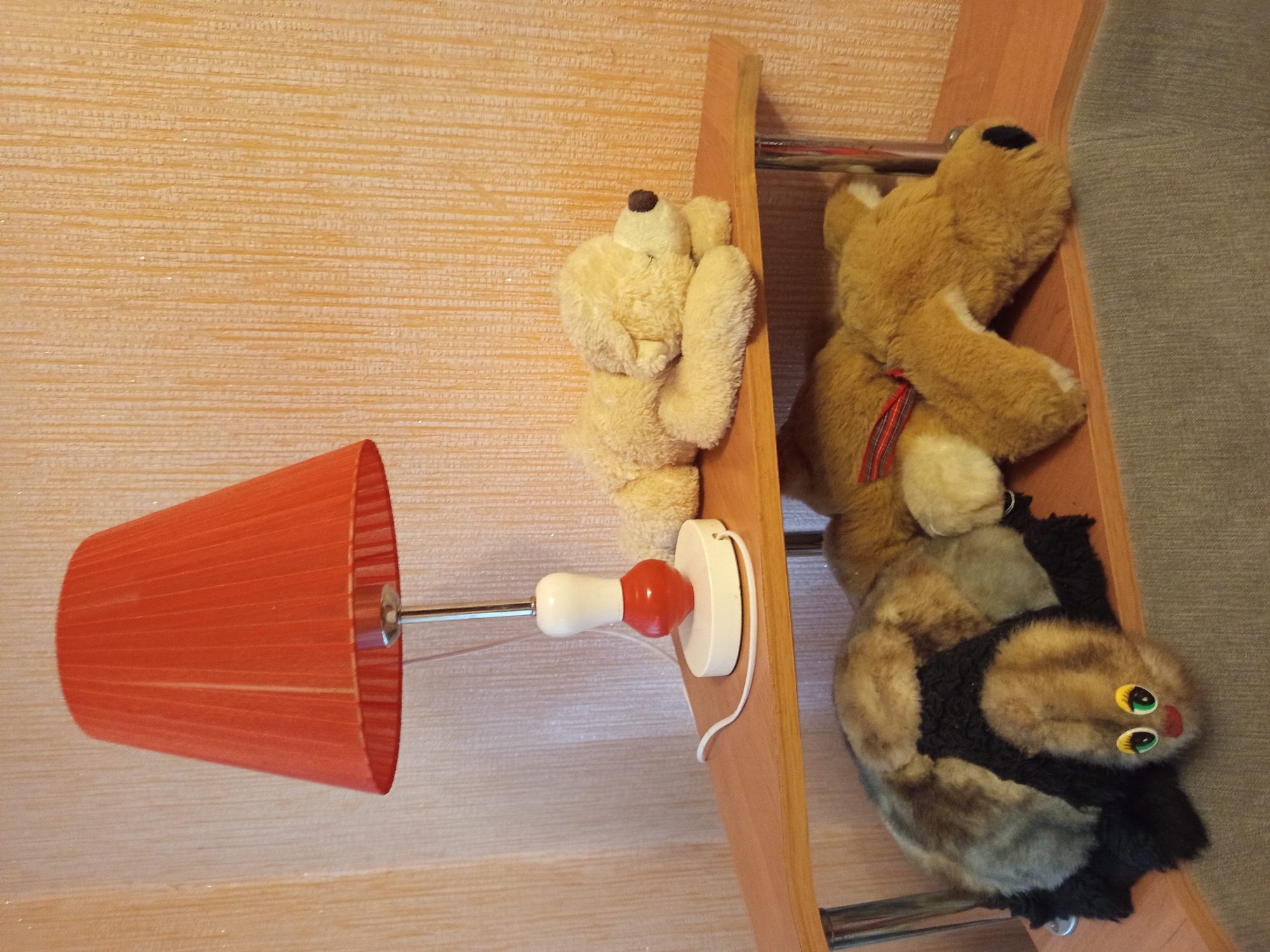 The image shows a collection of stuffed animals on a wooden shelf against a textured, light-brown wall. On the left, a light-colored teddy bear is propped up against the shelf. To its right, a larger brown teddy bear with a red and black striped ribbon around its neck is also resting on the shelf. Below the brown teddy bear, a black and brown stuffed animal, resembling a cat with large, round eyes, is positioned. A red lampshade with a metal stem and a white and red knob is attached to the wall on the left side of the image. The scene appears to be indoors, possibly in a child's room or a decorative display. The lighting suggests it could be daytime, with no indication of specific weather conditions. There is no visible text in the image. The overall impression is one of a cozy and playful arrangement of toys.