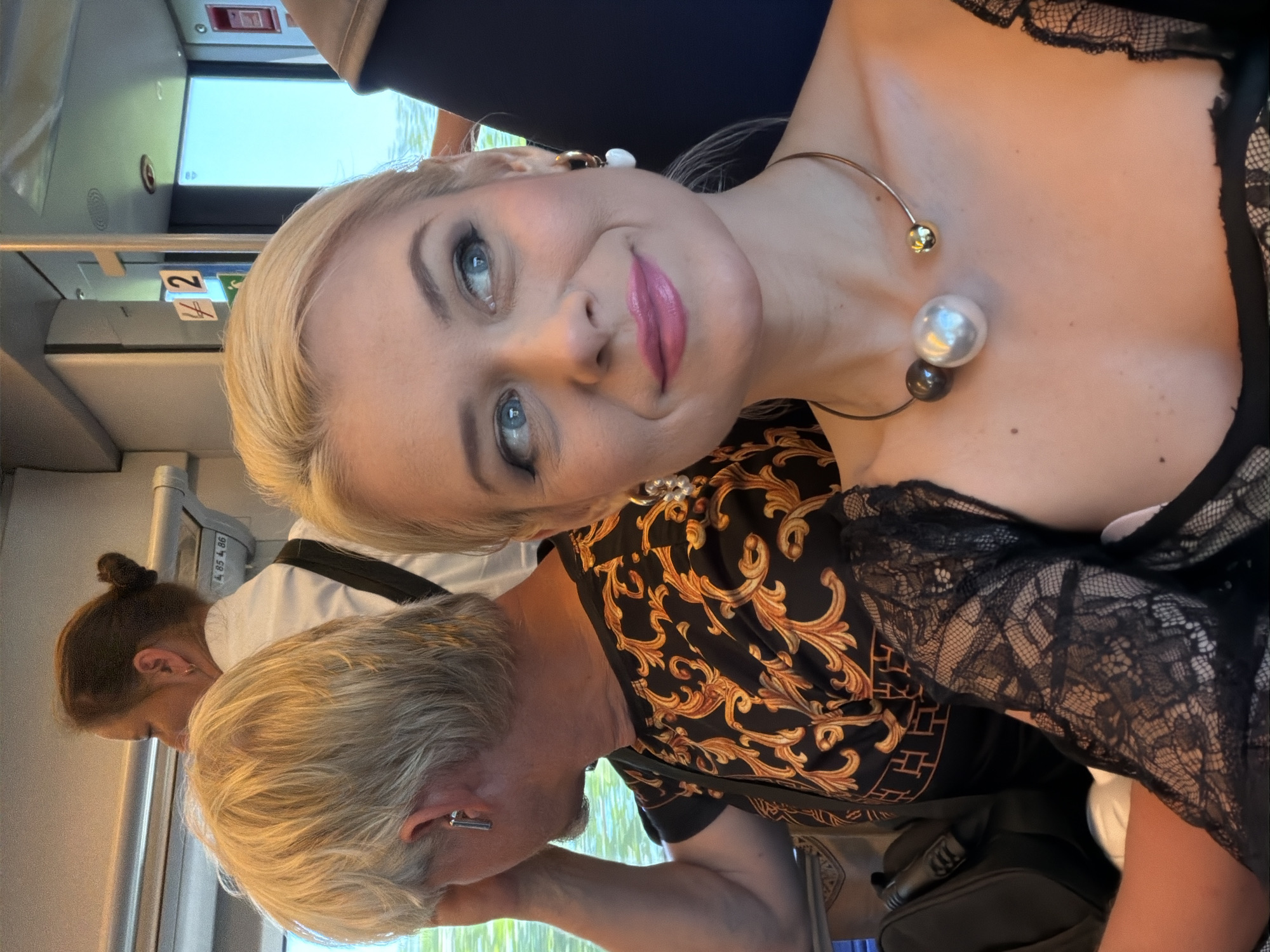 In Sochi, Russia, a blonde woman with blue eyes is seen from the chest up, looking to her right with a slight smile. She is wearing a black lace top, a statement necklace with three large spherical beads of varying colors (dark gray, white, and gold), and white heart-shaped earrings. Next to her, a person with blond hair is partially visible, wearing a black shirt with an ornate gold pattern. Behind them, another individual with dark hair pulled into a bun is seen from the shoulder up, wearing a white shirt and a black strap. The scene appears to be inside a train car, indicated by the presence of windows showing a blurry outdoor scene and signage on the wall, including numbers and symbols. The lighting suggests it is daytime.
