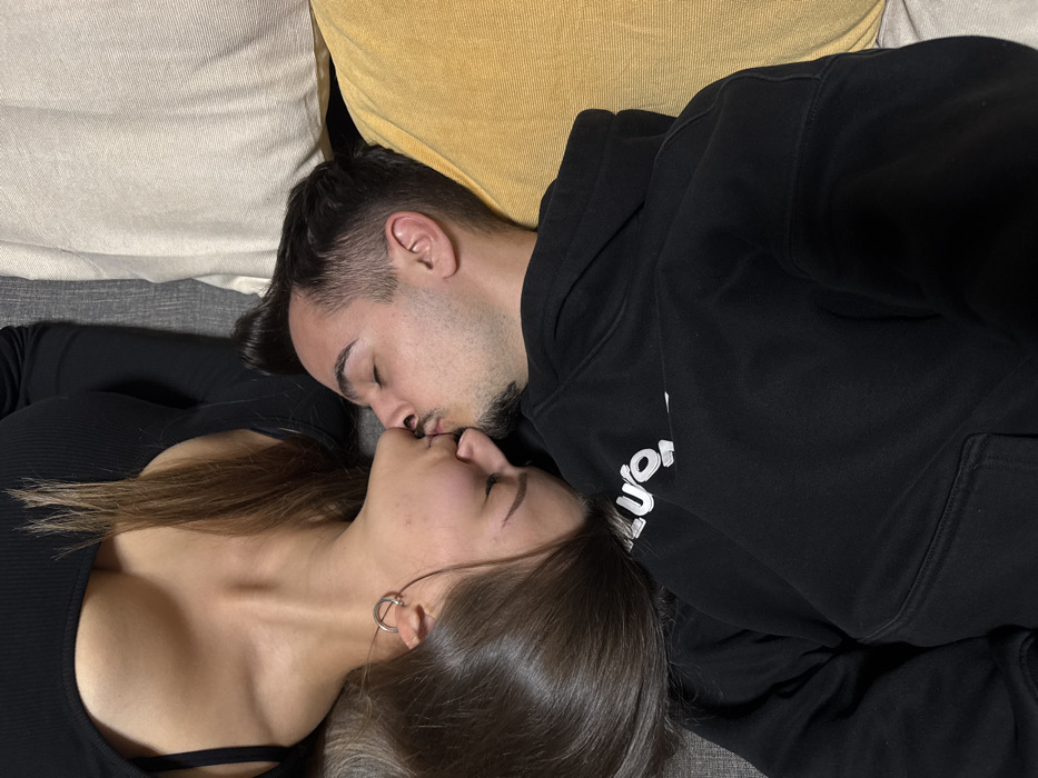Two individuals, a female and a male, are depicted in an intimate indoor setting within the city of Sofia, Bulgaria. The female, with long dark brown hair and wearing a black ribbed long-sleeved top, lies horizontally, positioned above the male. She has a silver hoop earring in her visible ear. The male, with short dark hair and facial stubble, lies horizontally on his back, wearing a black hooded sweatshirt with partial white lettering visible on the left shoulder. Both individuals are kissing mouth-to-mouth, with their eyes closed. They are resting on a grey upholstered surface, flanked by a cream-colored cushion and a yellow cushion to the male's right.
