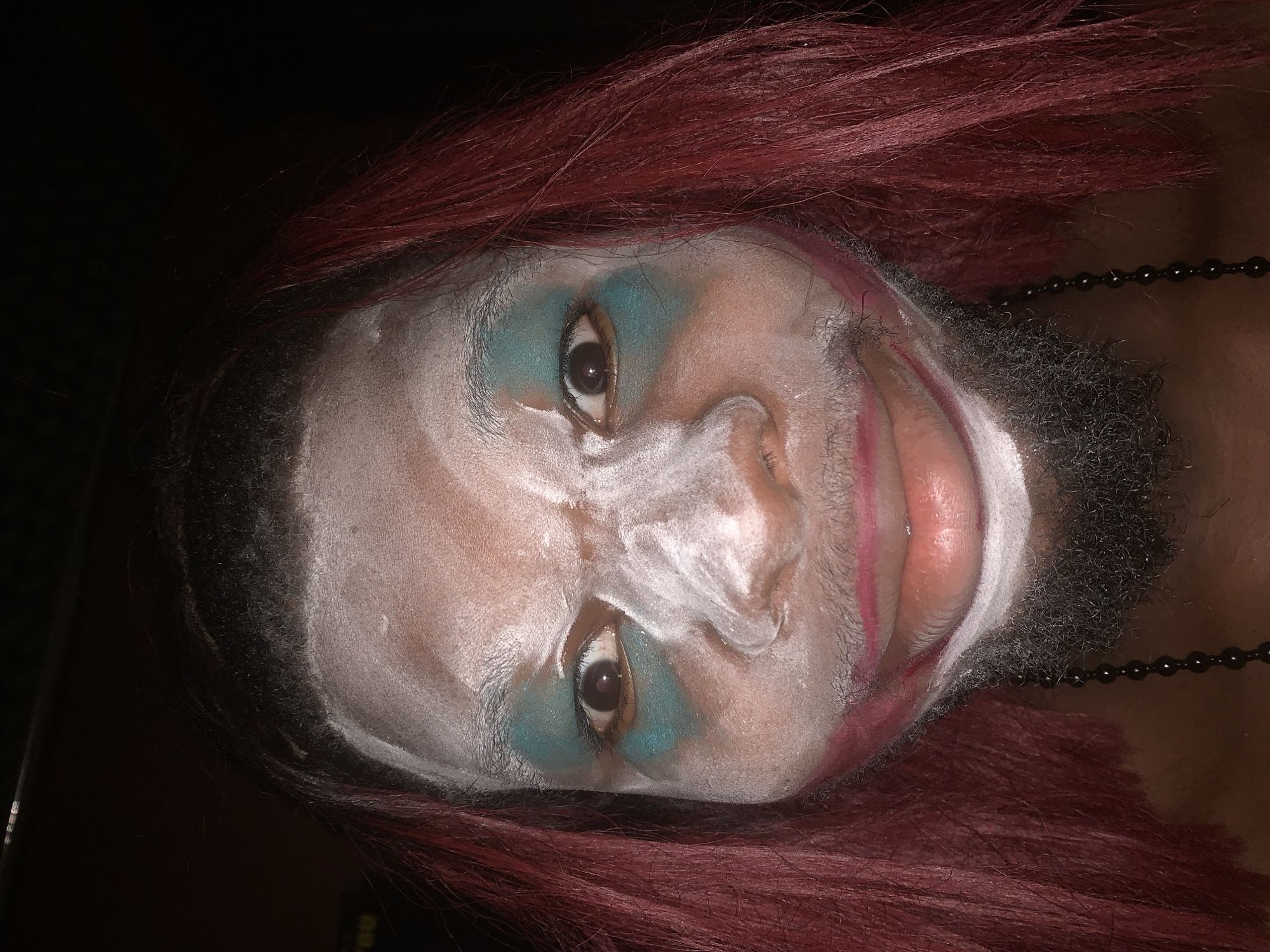 The image displays a close-up portrait of a person with dark skin, oriented sideways. The individual wears elaborate makeup, featuring prominent white face paint covering large portions of their forehead, nose, cheeks, and chin, creating a theatrical or clown-like appearance. Teal or turquoise eyeshadow is heavily applied around both eyes, extending outwards. Faint pink or red lines are visible on the cheeks and around the mouth, suggesting further makeup details or artistic smudging.
The person has dark facial hair, likely a beard, on their chin and jawline, some of which is covered by the white paint. They have dark reddish-brown hair, possibly a wig, framing their face and falling around their head. Both eyes are open, with dark brown irises gazing directly towards the viewer. The lips appear natural or lightly made up, with some red liner visible.
A black beaded necklace is visible on the person's neck and shoulder, partially hidden by their hair. The background is uniformly dark and indistinct, suggesting low light conditions or a dark backdrop. The overall scene conveys a pose for the camera, highlighting the dramatic makeup application. There is no visible text in the image. The photo was taken in Nkpor, Nigeria.