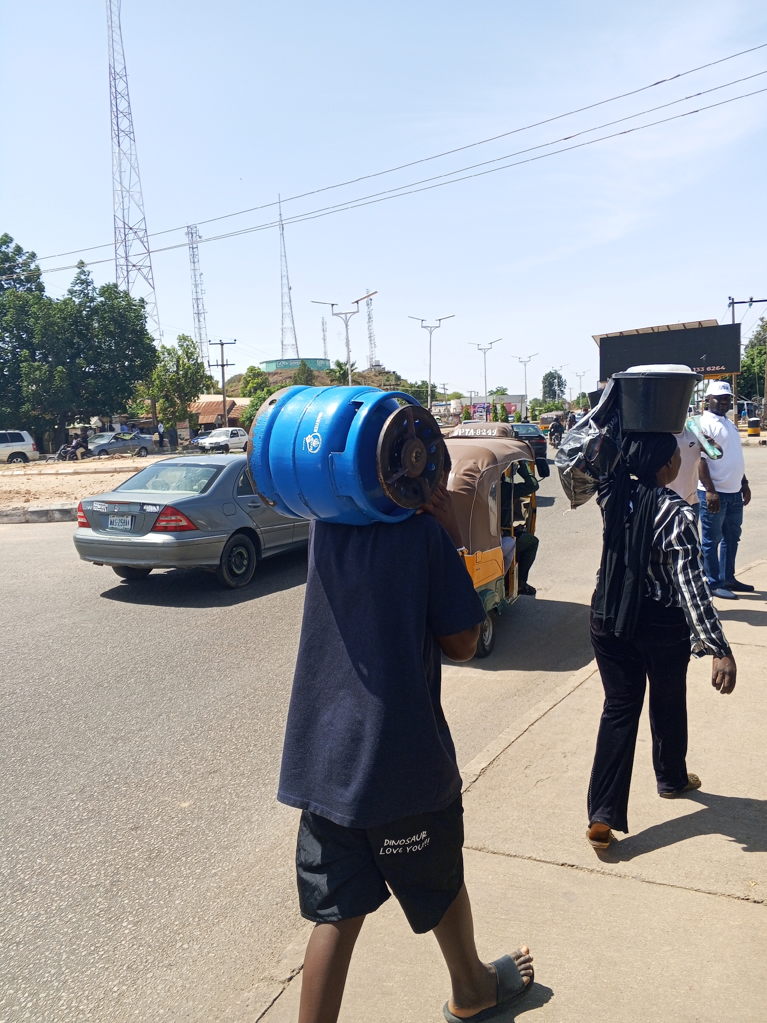 The image captures a vibrant street scene in Jos, Nigeria, under bright, clear daylight, casting long shadows. In the foreground, a person walks on a concrete sidewalk, seen from the waist down, wearing a dark blue t-shirt and black shorts. They are balancing a bright blue gas cylinder on their head. The shorts bear the white text "DINOSAUR LOVE YOU!!". This person is wearing black slide sandals.
To their right, slightly further along the sidewalk, another person is walking away from the viewer, carrying a black bucket or container on their head, along with other bundled items. This individual wears a black and white striped long-sleeved shirt and dark trousers. A brown and yellow motorized rickshaw, known as a Keke Napep, is visible in the middle ground, moving in the same direction as the pedestrians. Part of its license plate, "PTA-62J9", is discernible.
On the paved road to the left, traffic is moving. A silver Mercedes-Benz sedan, with the Nigerian license plate "NAS-258AA", is prominently visible. In the background, other vehicles, including cars and a white SUV, are parked or moving.
The scene features typical urban infrastructure: tall, lattice-style communication towers dominate the skyline in the distance, alongside utility poles and power lines crisscrossing the sky. Streetlights line the road, and green trees provide shade. A large, dark billboard with white text, including "133 6264", stands by the roadside further down. Hilly terrain with scattered buildings and structures can be seen in the background. The overall impression is one of daily life and commerce in an active urban environment.