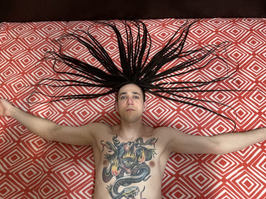 A person lies supine on a bedspread with a red and white diamond pattern. Their arms are outstretched to each side. The person has fair skin, short hair, and dreadlocks that fan out around their head. A large, colorful tattoo depicting a Medusa-like figure with snakes and an eye covers their chest. The person appears to be staring directly at the camera. The composition is a high-angle shot, emphasizing the symmetrical arrangement of the person's body and hair.