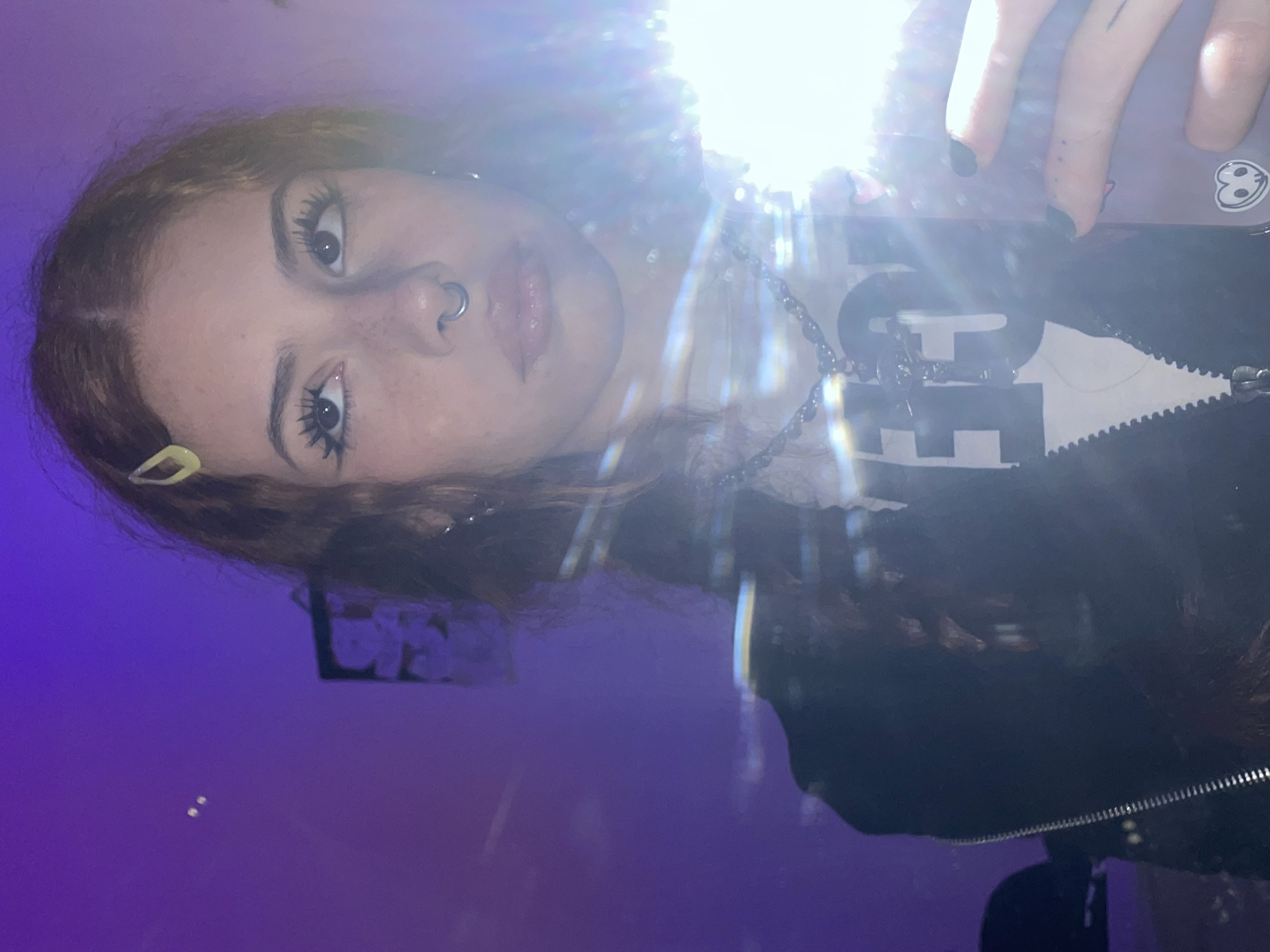 A young woman is taking a selfie in a room with purple walls. She is wearing a black zip-up jacket over a white shirt with a graphic design. She has a nose ring, a necklace, and a yellow hair clip. Her hair is light brown and wavy. The flash from her phone is very bright. A picture frame is on the wall behind her. It is not possible to determine the location in Buenos Aires, Argentina from this image.