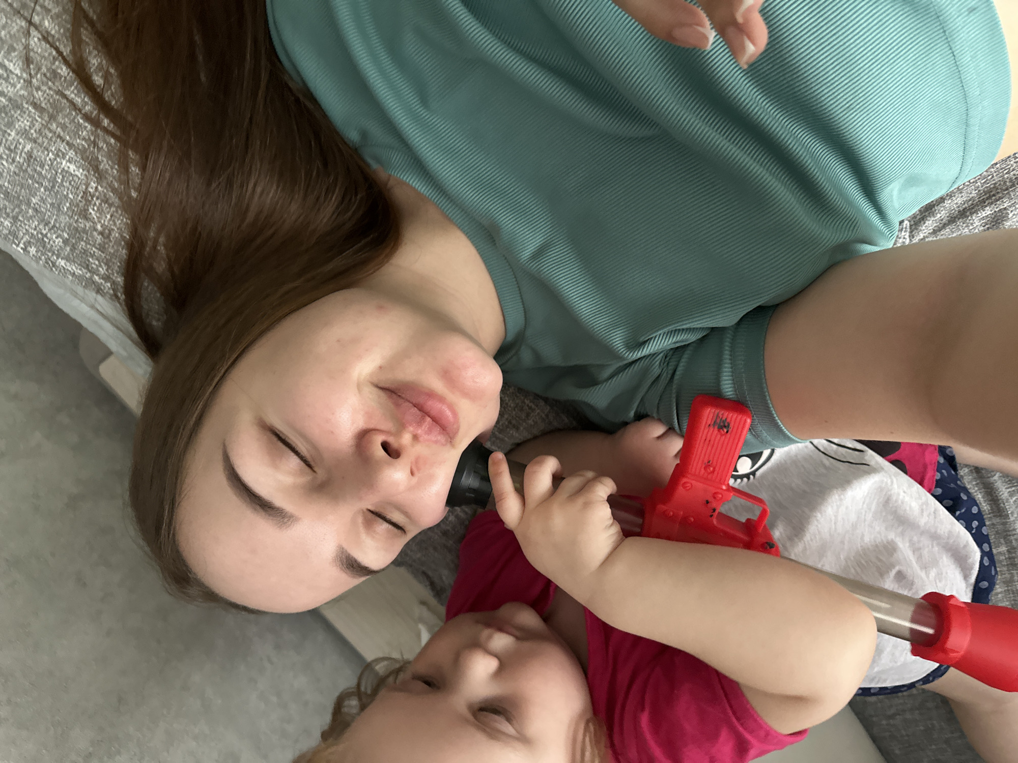 A woman with long brown hair and closed eyes is lying on a gray textured surface next to a child. The child is wearing a pink shirt and holding a red toy. The woman is wearing a teal top. Part of the child's patterned clothing is also visible. No specific details indicate the location is Chaikovsky, Russia.