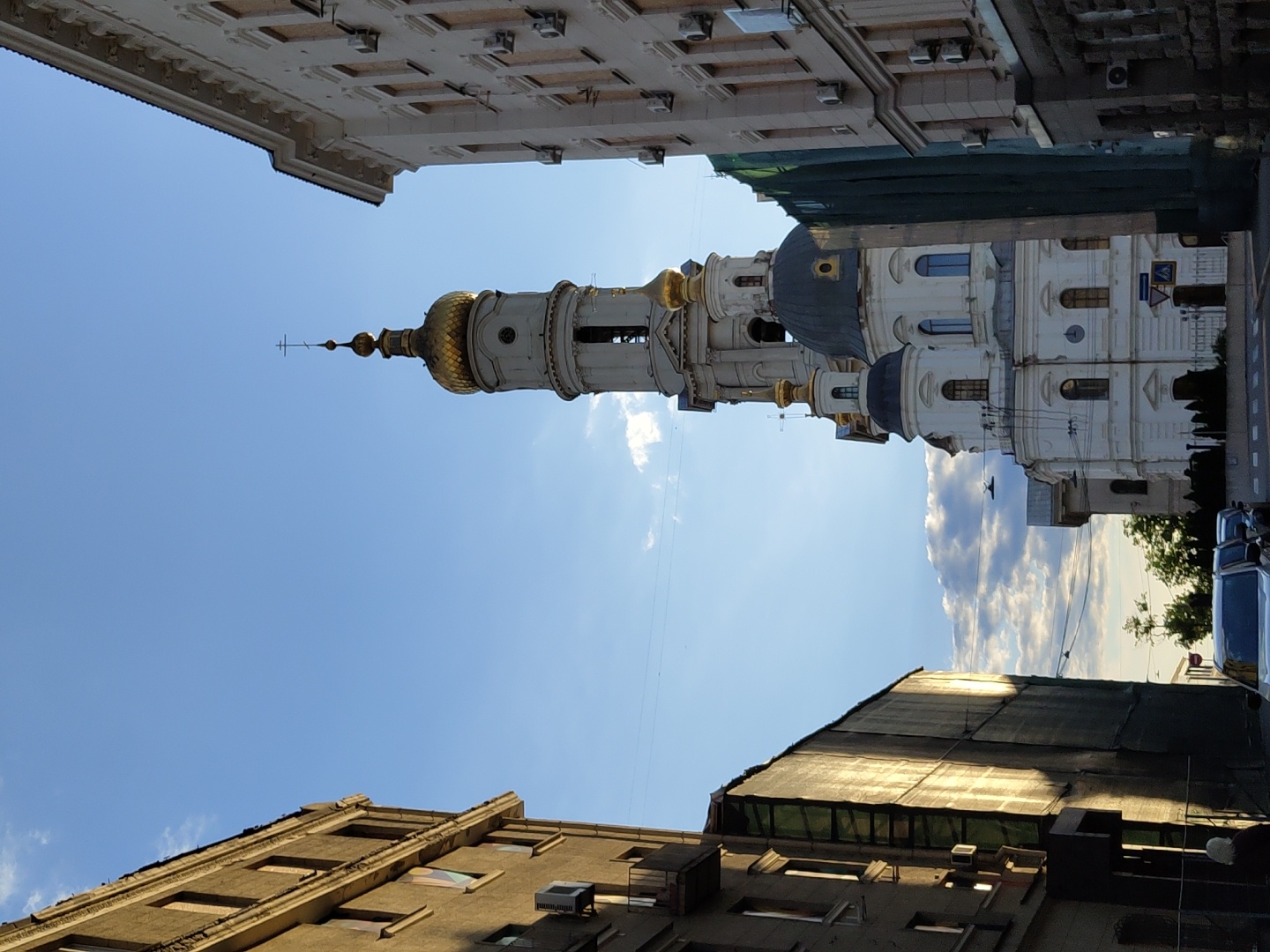 A street-level photograph captures a tall, ornate Orthodox Christian church or cathedral against a bright blue sky. The main structure is predominantly white, featuring multiple golden onion domes and several smaller dark blue or grey domes. A prominent, slender bell tower or spire, also white with a golden cupola, rises centrally, topped with a distinctive Orthodox cross.
The church is flanked by other urban buildings. On the right, a modern-looking, light-colored building with multiple windows can be seen. On the left, an older, darker-toned building with a noticeable roofline is partially visible. Behind the church, to its left, part of another building appears to be undergoing renovation, covered in green scaffolding netting.
The sky is a vibrant blue with scattered white cumulus clouds, indicating clear weather. The lighting suggests it is late morning or early afternoon, with the sun high above. In the lower right corner, a few parked cars are faintly visible on what appears to be a street, along with a blurry blue and white street sign. No people are present, and no specific activity is taking place. The scene conveys a sense of architectural grandeur and urban tranquility under a pleasant sky.