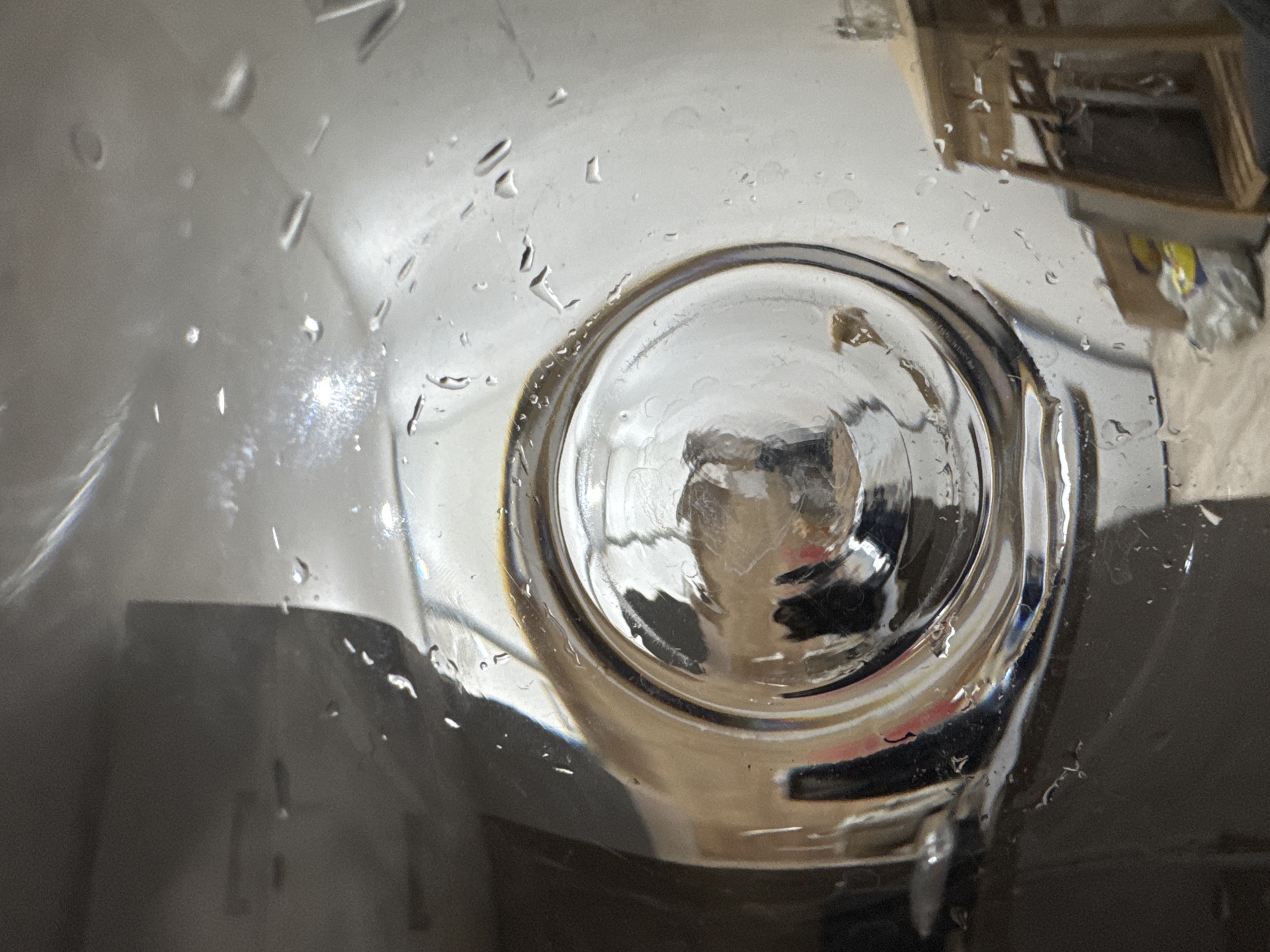 The image shows a close-up, low-angle view through a glass container, possibly a jar or a bottle, which has water droplets on its interior surface. The container is reflecting its surroundings. The most prominent feature is the concave bottom of the container, seen from the inside, which acts as a lens distorting the reflected image.
The reflection within the container reveals a dimly lit interior space. We can discern shapes that suggest furniture or objects, including what appears to be a light-colored, possibly wooden, shelving unit or table on the right side of the frame. In front of this structure, there are some items on the floor, including what looks like a plastic bag and possibly some cardboard. The background walls are a light, neutral color, and the floor appears to be a light-colored laminate or tile.
The lighting is ambient, with some areas brighter than others, creating reflections and highlights on the wet glass. The overall impression is that of an indoor setting, perhaps a home or storage area, viewed from within a glass vessel.
Regarding the location in Trenčín, Slovakia, this image provides no specific visual cues to identify its precise location within the city. The contents of the reflection are generic indoor elements that could be found in many residential or commercial spaces. Therefore, it is impossible to determine the specific location in Trenčín based solely on this image.
