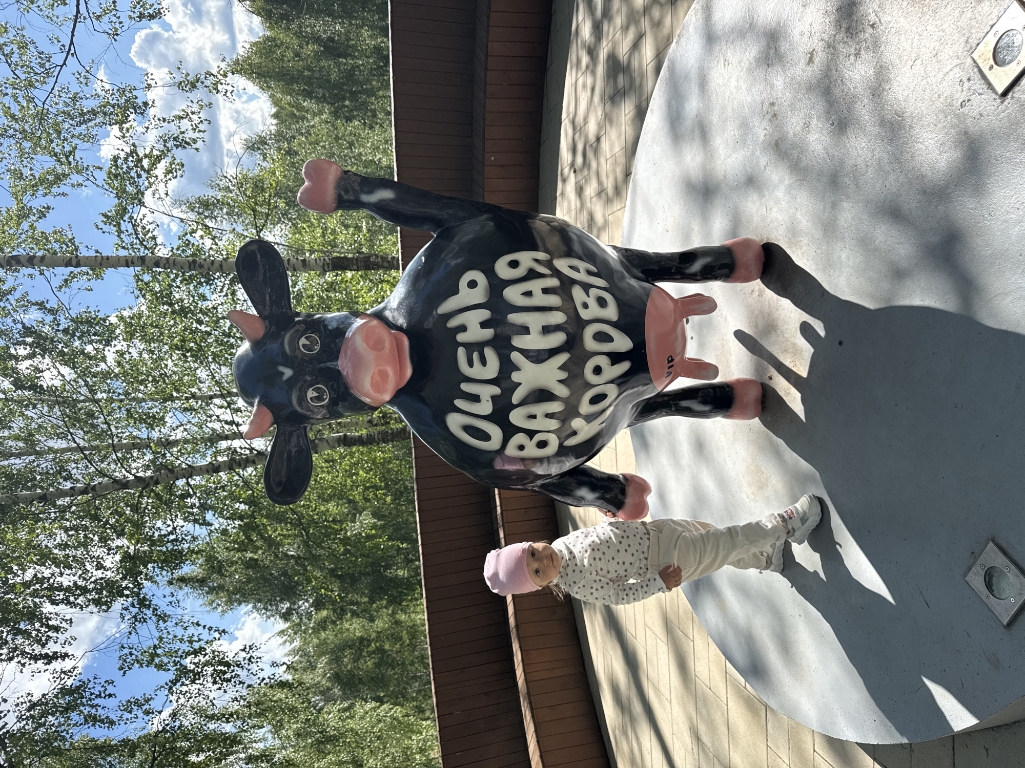 A black-and-pink cow statue, inscribed with Russian text, stands on a circular platform in an outdoor setting. A small child in white clothing and a pink hat is positioned next to one of the cow’s feet. Behind the statue, a fence and trees suggest a park or similar environment. The location is Zelenodol'sk, Russia.