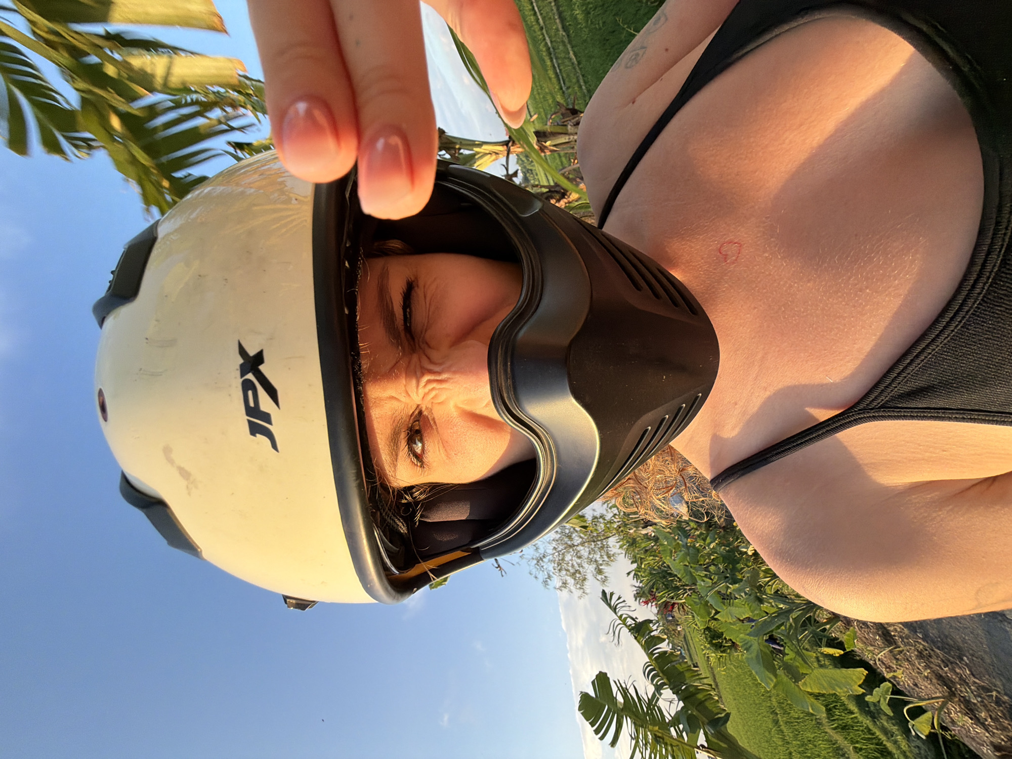 A close-up, low-angle selfie features a woman wearing a white open-face helmet with a black chin guard and a black tank top. She is outdoors, with a bright blue sky above and a lush green rice paddy field and tropical foliage in the background. The lighting suggests it is either sunrise or sunset, casting a warm glow on her skin. Her face is partially obscured by the helmet, and she is squinting and making a playful expression, with her right hand held up to the camera, fingers extended and obscuring part of the frame. A small, red heart tattoo is visible on her chest. The location is specified as North Kuta, Indonesia, a region known for its scenic rice terraces and agricultural landscapes.