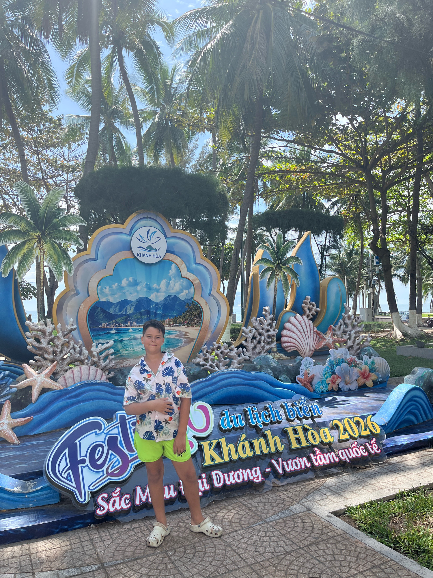 A young boy stands in front of a large, colorful beach-themed display. The display features a backdrop of a blue seashell shape with a scenic view of mountains, a beach, and the ocean. It also includes decorative elements like coral, seashells, and starfish, as well as large lettering that reads "du lịch biển Khánh Hòa 2026" and "Sắc Màu Dưỡng - Vươn Tầm Quốc Tế," which translates to "Beach Tourism Khanh Hoa 2026" and "Colorful Journey - Reaching International Level." The boy is wearing a white short-sleeved shirt with a blue floral pattern and bright green shorts. He is also wearing white clogs. The scene is set outdoors with numerous palm trees and other foliage visible in the background. This is a promotional display for the Khanh Hoa province beach tourism event in 2026, likely located in the city of Nha Trang, Vietnam.