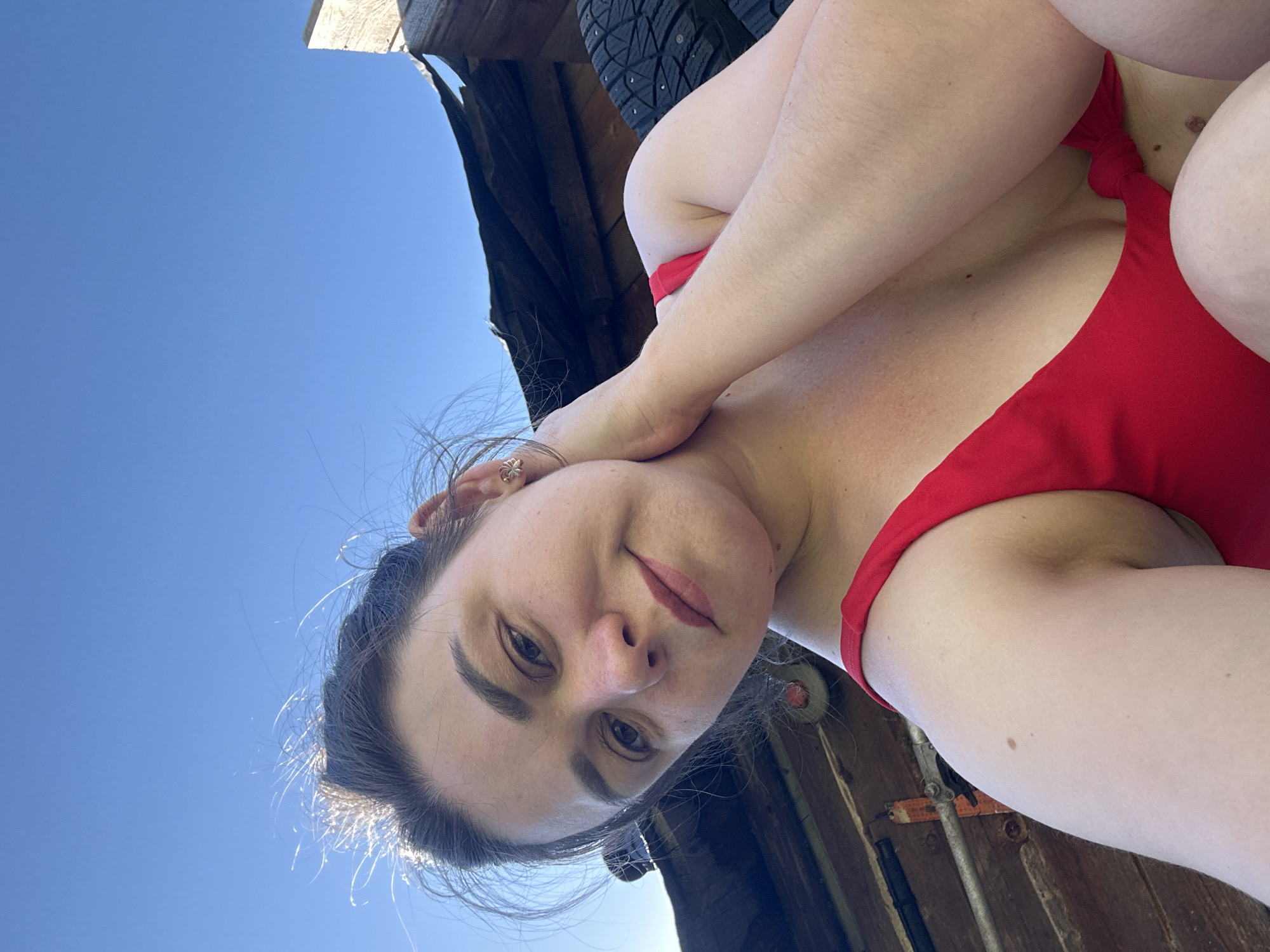 A woman is wearing a red swimsuit in what appears to be an outdoor setting in Chaikovsky, Russia. She is partially under a wooden structure that may be a shed or building overhang, with a blue sky visible above. A tire with studs is visible behind her.