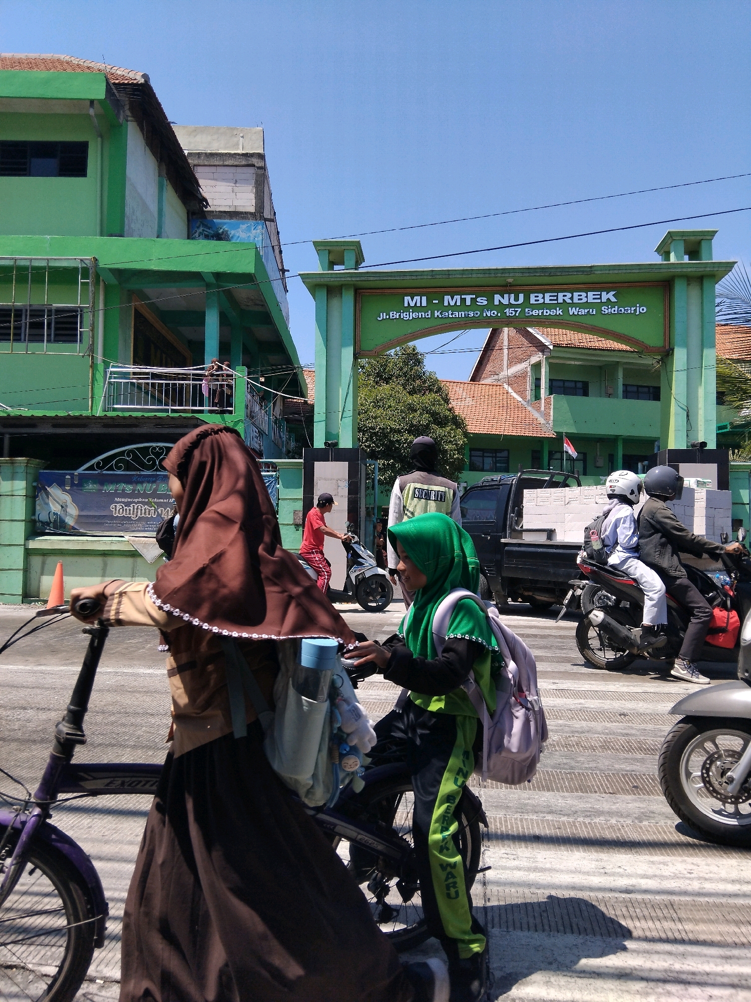 In the city of Kepuh Kiriman, Indonesia, a sunny day unfolds with vibrant activity outside a green-hued educational institution. The building, identified by the signage "MI - MTs NU BERBEK" with the address "Jl. Brigjend Katamso No. 157 Berbek Waru Sidoarjo", features multiple levels and an arched entranceway.
In the foreground, a young individual on a purple bicycle, wearing a brown hijab and a dark brown skirt, is partially obscured. They are carrying a light blue water bottle and a light blue bag with stuffed animal charms. To their right, another young person in a bright green hijab and matching tracksuit, with a light gray backpack, is also on a bicycle.
The street is busy with traffic. A black pickup truck, laden with white bricks, is parked on the right side of the road. A motorcycle carrying two passengers, both wearing helmets, passes by the truck. The passenger in the back wears a white top and gray pants, while the driver is in a dark jacket. Further back, another motorcycle with a rider in a red shirt and checkered pants is visible.
The environment is characterized by clear blue skies and the architectural style of the school building, which includes green painted walls, white accents, and a tiled roof. The road surface appears to be paved with distinct lines, possibly indicating a pedestrian crossing. The overall scene captures a moment of daily life and transportation within an Indonesian community.
