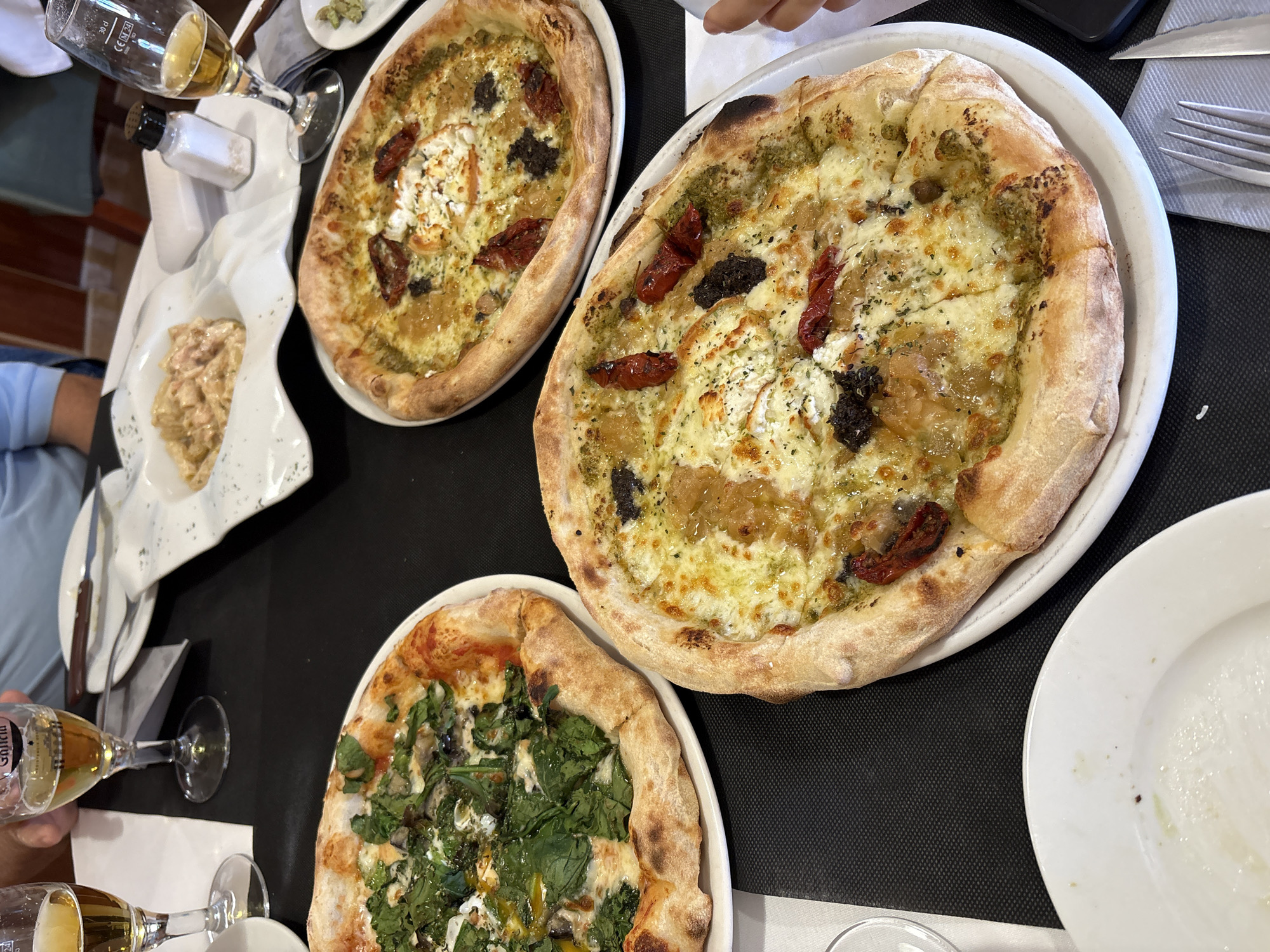 The image presents an overhead view of a dining table in what appears to be a restaurant or pizzeria in Teulada, Spain, where several people are enjoying a meal. Three pizzas are the central subjects, each on a white plate.
Two of the pizzas share similar toppings: a vibrant green base, possibly pesto, covered with melted white cheese, chunks of crumbled white cheese (likely feta or goat cheese), sun-dried tomato pieces, and dark, irregular elements that could be olive tapenade or mushrooms. A glossy, amber-colored topping, possibly caramelized onions or a sweet drizzle, is also visible. Both feature a thick, slightly charred crust.
The third pizza, partially in view in the foreground, is topped with abundant fresh green leaves, likely spinach, melted cheese, and what appears to be mushrooms and a distinctive bright yellow element, possibly an egg yolk or a rich sauce.
The table is set with black placemats. Several glasses are visible, two of which contain a light amber beverage, suggesting beer or white wine. The text "GALICIA" is clearly visible on one of these glasses. A white salt shaker with a black cap, a small white bowl containing a creamy side dish (perhaps a pasta salad), and cutlery (forks and a knife) resting on a grey napkin complete the setting. Hands and forearms of at least two diners are visible at the edges of the frame, indicating an ongoing meal. The overall atmosphere is casual and convivial.