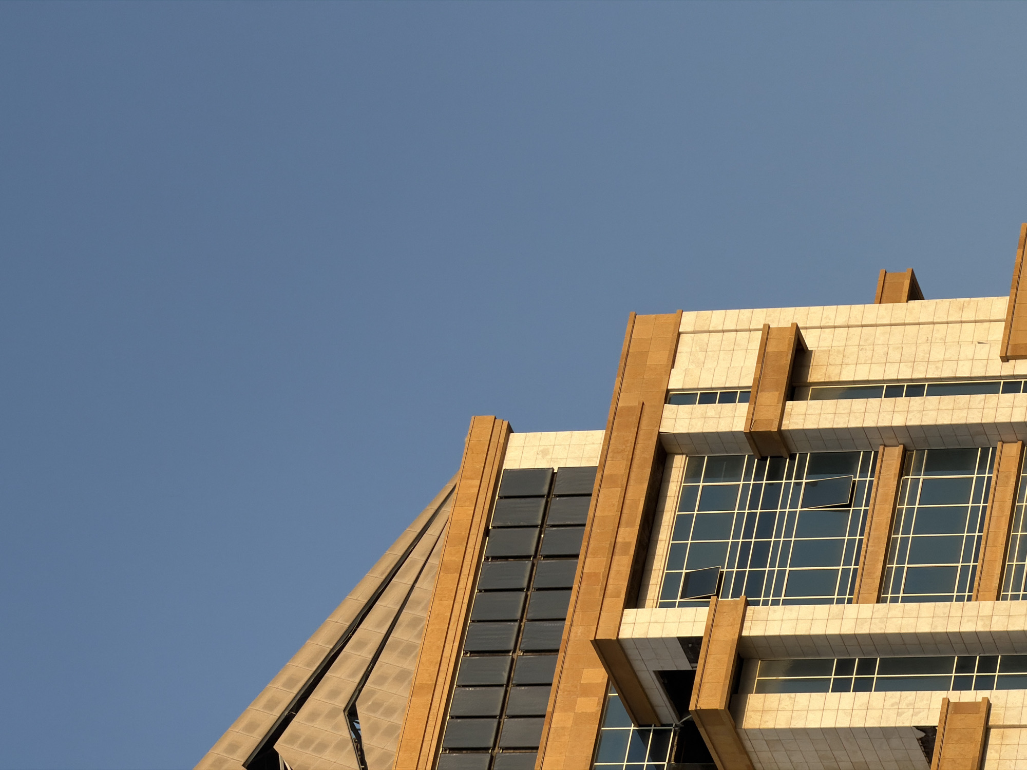 The image captures a close-up, angled view of a modern building with a tiered, stepped design, set against a clear, bright blue sky. The building's facade is a combination of light-colored stone tiles and dark-tinted windows, with prominent, horizontal bands of golden-brown material accentuating the different levels. Several windows are visible, some with panes opened, revealing the interior structure. The top of the building features a distinctive angled roofline with a textured surface, also in a golden-brown hue. The lighting suggests it is daytime, with the sun casting a warm glow on the building's surfaces, highlighting its architectural details. The image is devoid of any people or other elements, focusing solely on the building's structure and its interaction with the sky.