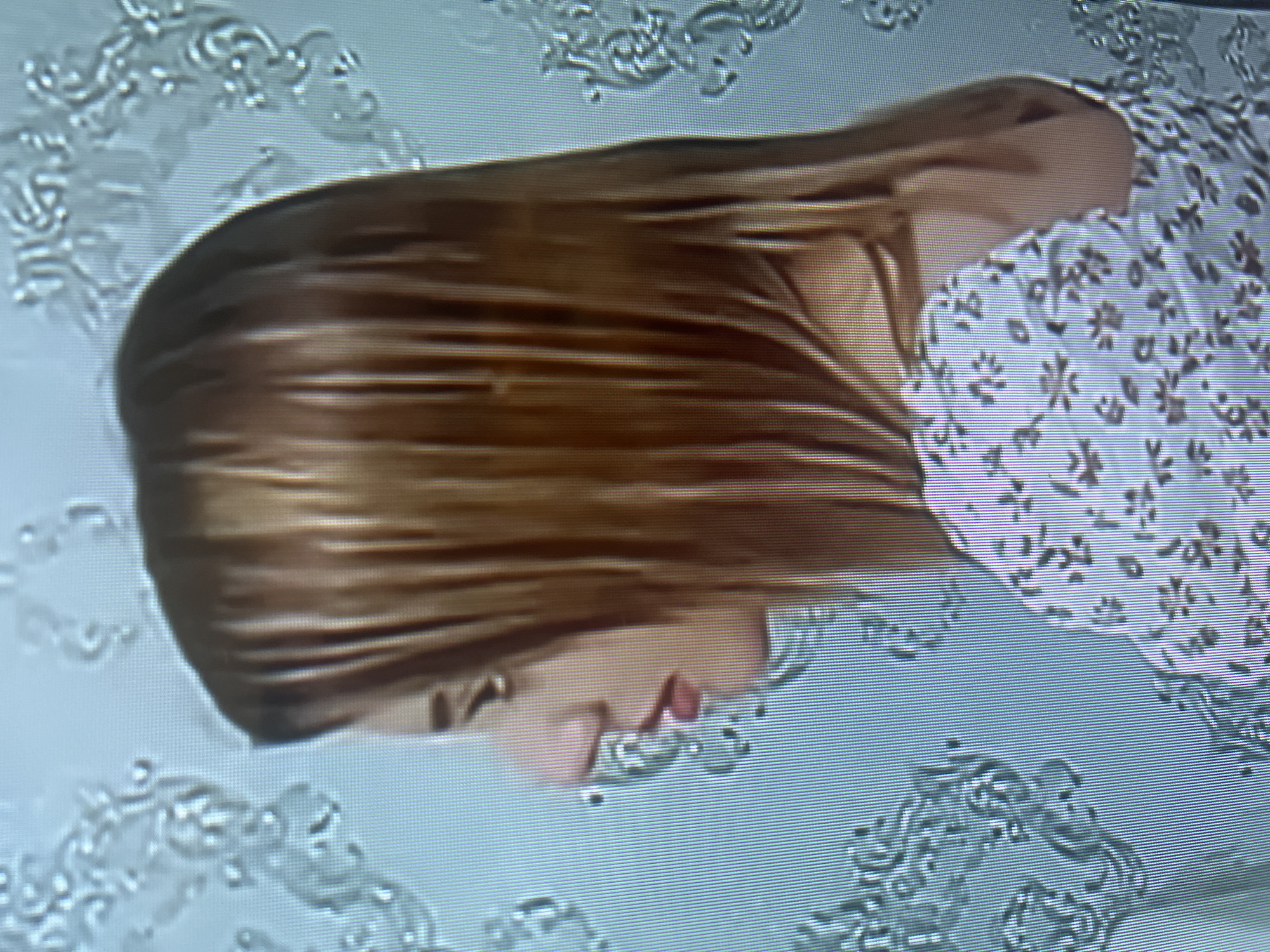 A profile view of a young woman is captured, facing left. She has light skin and her head is turned, revealing a prominent nose and lips with red lipstick. Her hair is shoulder-length and appears to be a warm brown with blonde highlights. She is wearing a white top with a floral pattern in muted tones. The background consists of a pale blue surface with a light, ornate pattern resembling wallpaper or a textile. The image quality is low, with visible pixelation and scan lines, suggesting it may be a screenshot from a video or a low-resolution photograph. The setting appears to be indoors, possibly a domestic environment, though no specific architectural details or urban context are discernible to place it within Osh City, Kyrgyzstan.