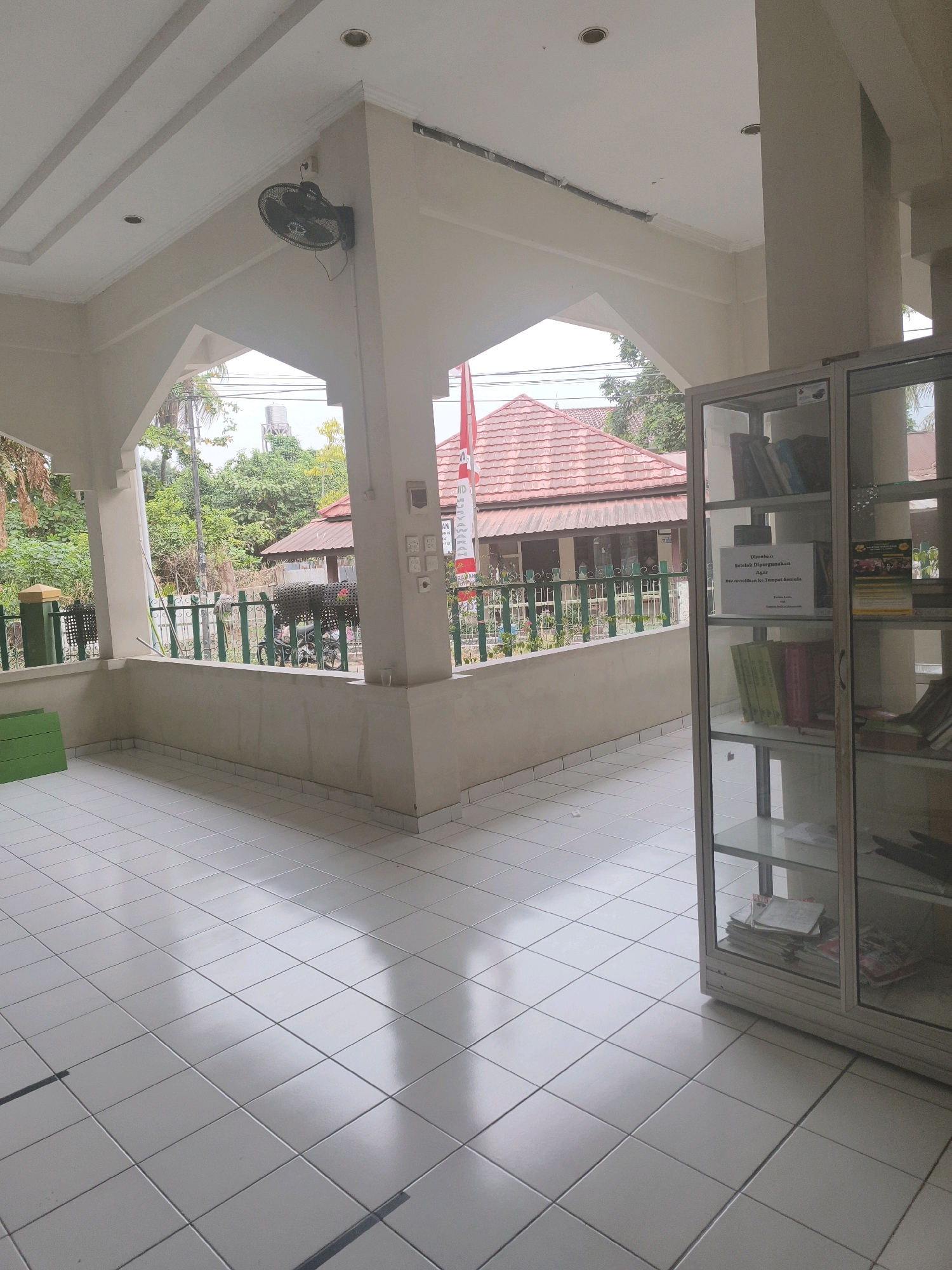 The image shows the interior of a covered outdoor area with a white tiled floor. In the foreground, to the right, is a glass-fronted display cabinet filled with books and papers. The cabinet is silver-framed and has multiple shelves. Beyond the cabinet, through an archway, is a view of trees, a red-tiled roof of a building, and a water tower in the distance. A red and white flag is visible on a pole outside. To the left of the archway, a black oscillating fan is mounted on a white wall. A green rectangular object, possibly a crate or bench, is visible against the wall on the far left. The scene suggests an outdoor seating or waiting area, possibly associated with a public building or institution in a tropical or subtropical climate.