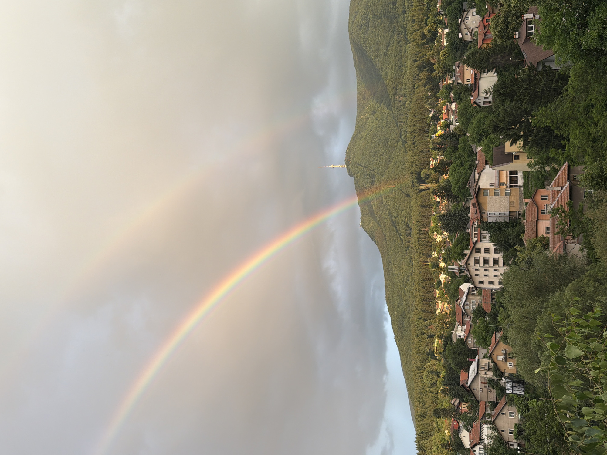 The image captures a scenic view of Sofia, Bulgaria, featuring a double rainbow arching across a cloudy sky. In the foreground, a dense collection of residential buildings with terracotta roofs is nestled on a hillside covered in lush green trees. Beyond the houses, a verdant, tree-covered mountain rises, crowned by a tall radio tower. The scene suggests a post-rain setting, with the vibrant colors of the rainbow contrasting with the moody sky and the peaceful, built environment. No people are visible in the image.