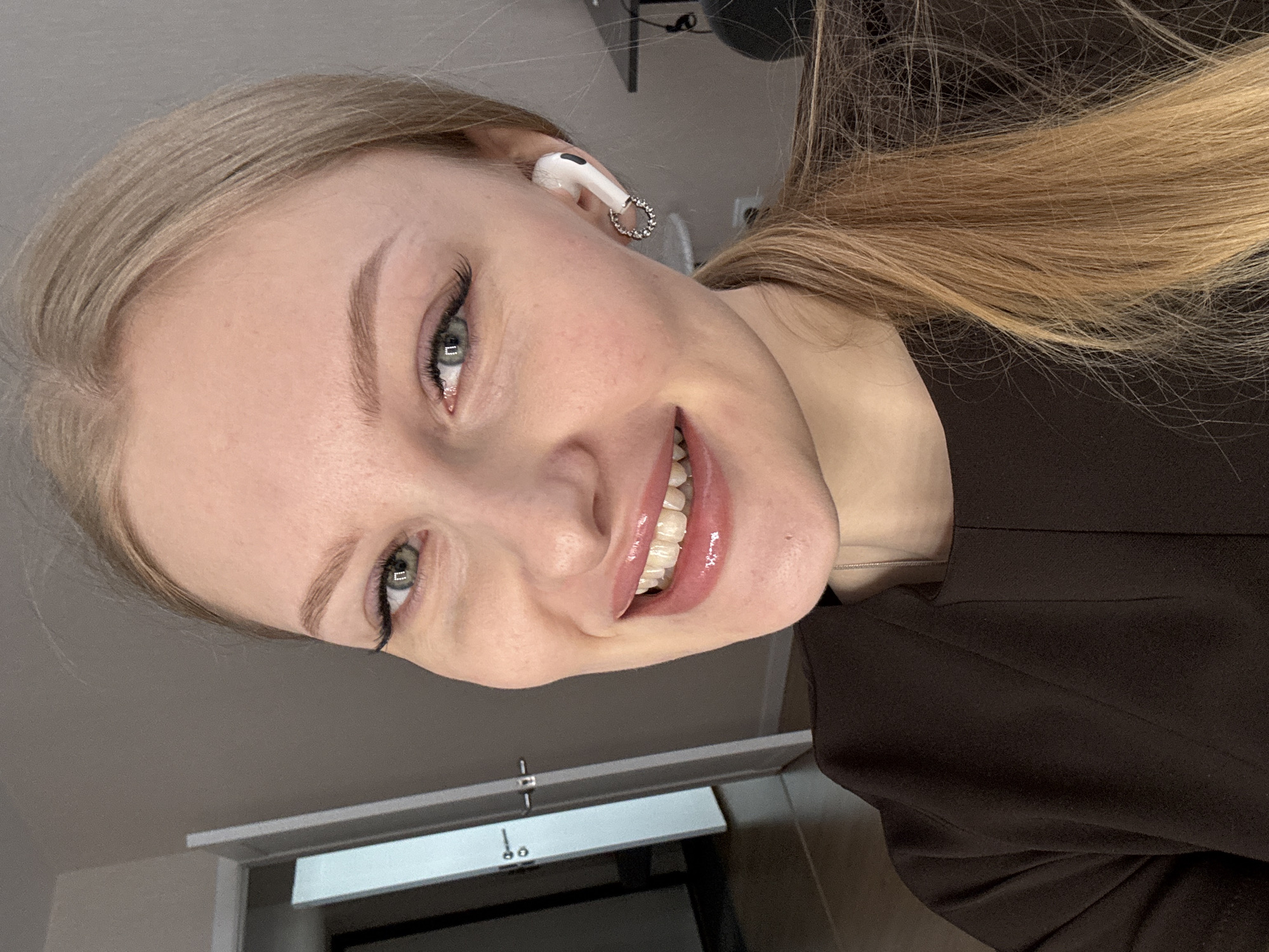 A close-up, slightly angled shot depicts a smiling young woman with fair skin, blonde hair pulled back, and light green eyes. She is wearing a white earbud with a dangling, circular charm. Her lips are glossy, and she appears to be wearing light makeup. She is wearing a dark brown top or jacket. The background includes a portion of a grey wall and a door frame. The location in Perm, Russia, cannot be determined from the image.
