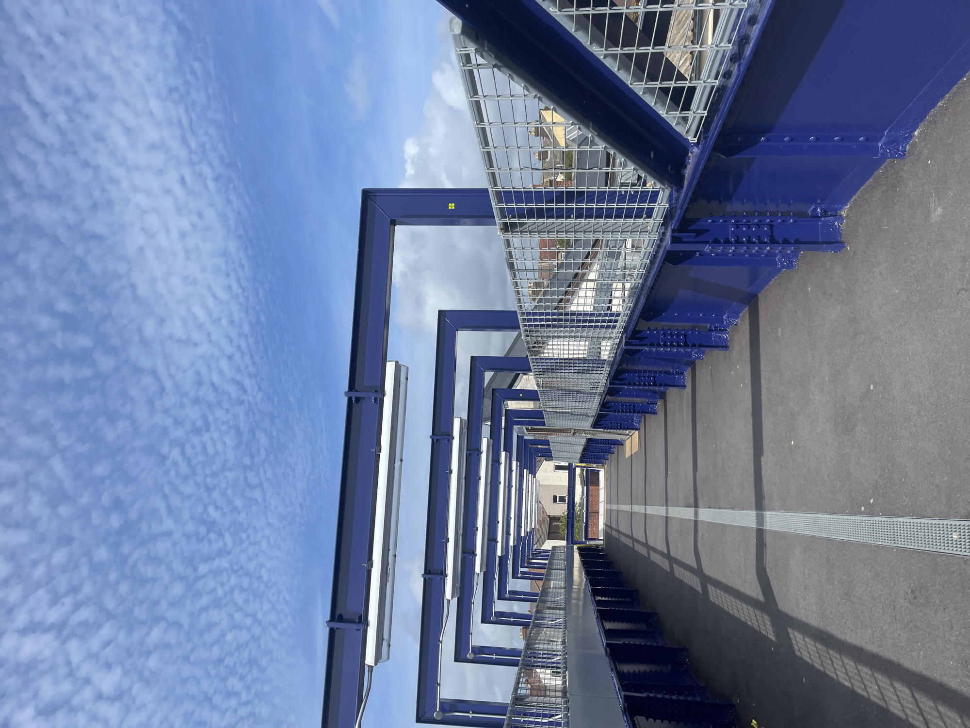 The image depicts an elevated pedestrian walkway or bridge, viewed from a low angle, looking forward along its length. When oriented correctly, the scene is bathed in bright daylight under a clear blue sky sparsely dotted with small, rippled clouds.
The most prominent features are a series of royal blue metallic structural beams that form a repeating archway or frame over the walkway. These robust beams are bolted together with visible rivets and extend into the distance, creating a sense of perspective. On the right side of the walkway, a silver metallic mesh railing runs parallel to the path, affixed to the blue structure. On the left side, several long, white fluorescent light fixtures are suspended from the blue overhead beams.
The walkway surface is dark grey asphalt, featuring a distinct light grey tactile paving strip running down its center. Strong, elongated shadows from the overhead structure and the right-side railing are cast across the path, indicating bright sunlight. In the background, through the framework of the structure and above the railing, the rooftops and upper parts of various buildings are visible, suggesting an urban or semi-urban setting.
No people are visible, and there is no specific activity taking place. A small yellow square with the number "22" in black is affixed to one of the blue structural beams. The modern, industrial design of the structure points to contemporary infrastructure, likely a public thoroughfare such as a footbridge or platform access.