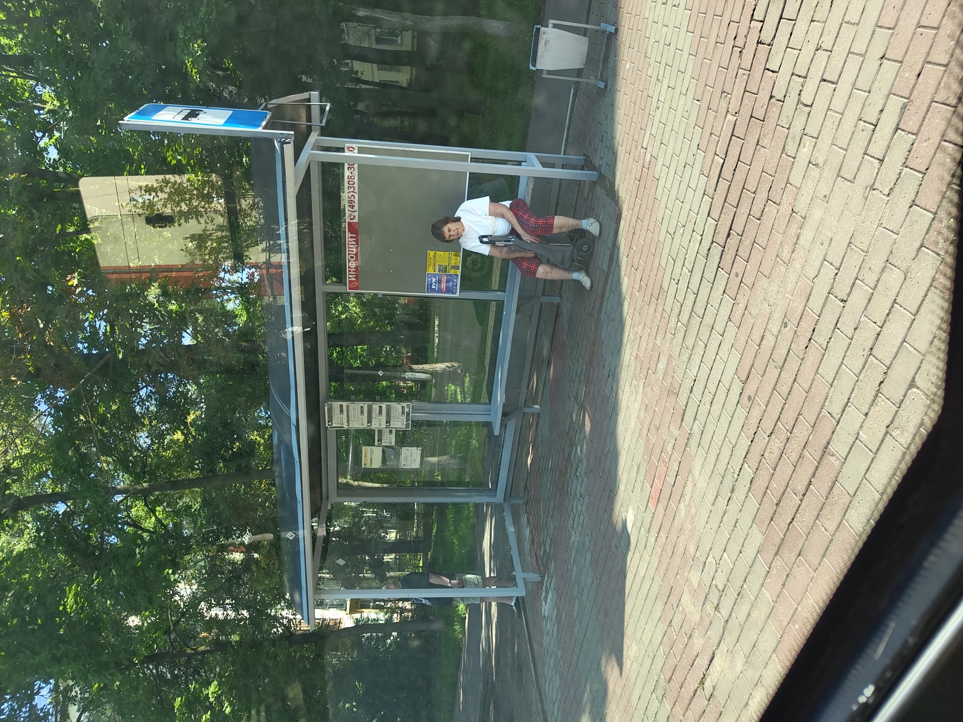 The image captures a street scene in Zhukovsky, Russia, featuring a bus stop shelter. A woman, seated on a bench inside the shelter, is the central human figure. She is wearing a white t-shirt and plaid pants and is holding onto the handle of a foldable shopping cart positioned in front of her. To her left, another person stands, partially obscured by the shelter's structure, wearing a dark shirt and shorts. The bus stop has a bench, a bulletin board with advertisements and schedules, and a trash can to the right of the shelter. The ground is paved with brick-like tiles. Lush green trees form a canopy behind the bus stop, suggesting a leafy urban environment. The day appears sunny, with shadows cast on the paved area.