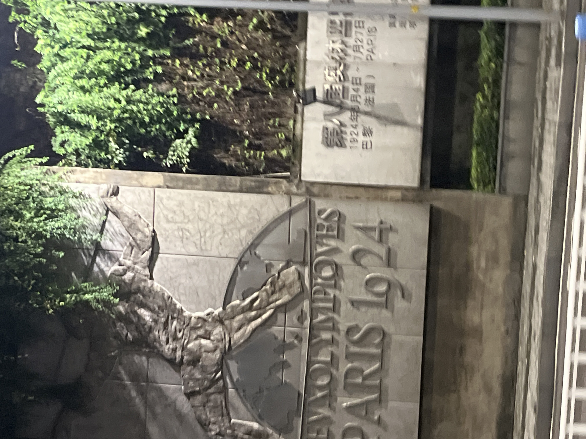 The image shows a relief sculpture on a wall, likely commemorating the VIII Olympiad held in Paris in 1924. The sculpture features stylized figures and text in both French and Chinese. The French text clearly reads "PARIS 1924" and likely includes "JEUX OLYMPIQUES" or a similar phrase. The Chinese text appears to refer to the same event, possibly with dates and location. The relief is weathered and has some plants growing around it. The background is blurred but suggests an outdoor urban environment, possibly a street at night with artificial lighting. The location is identified as the city of Jiulongpo District, China.