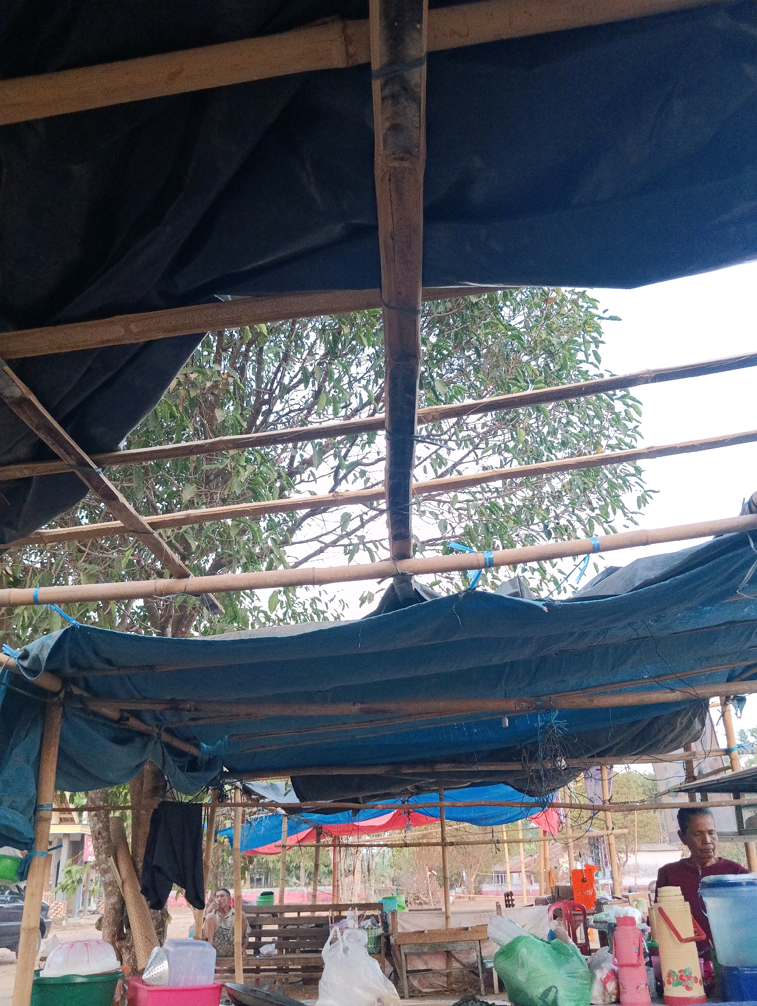 The image shows an outdoor market stall with a bamboo and tarp roof. A person wearing a maroon shirt is standing behind a table laden with various items, including thermoses, containers, and plastic bags. In the background, a wooden bench is visible, and another person is seated on it. The scene appears to be a casual, everyday market setting in Pematang Jaya, Indonesia, with trees and buildings visible in the distance. The overall atmosphere is rustic and informal.