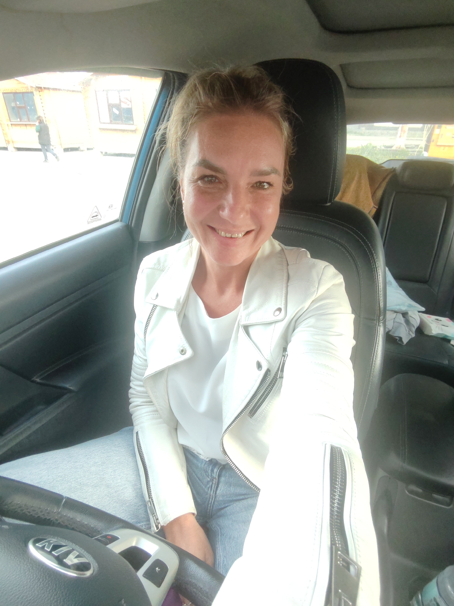 A woman is taking a selfie inside a car. She is smiling at the camera and wearing a white leather jacket over a white top and blue jeans. The car's interior, including the black leather seats and steering wheel, is visible. A person can be seen walking in the background through the car window, past some wooden buildings. The scene appears to be outdoors, and the lighting suggests it is daytime. The steering wheel has a "KIA" logo on it. There is a slight sheen on the woman's face, possibly from sweat or makeup.