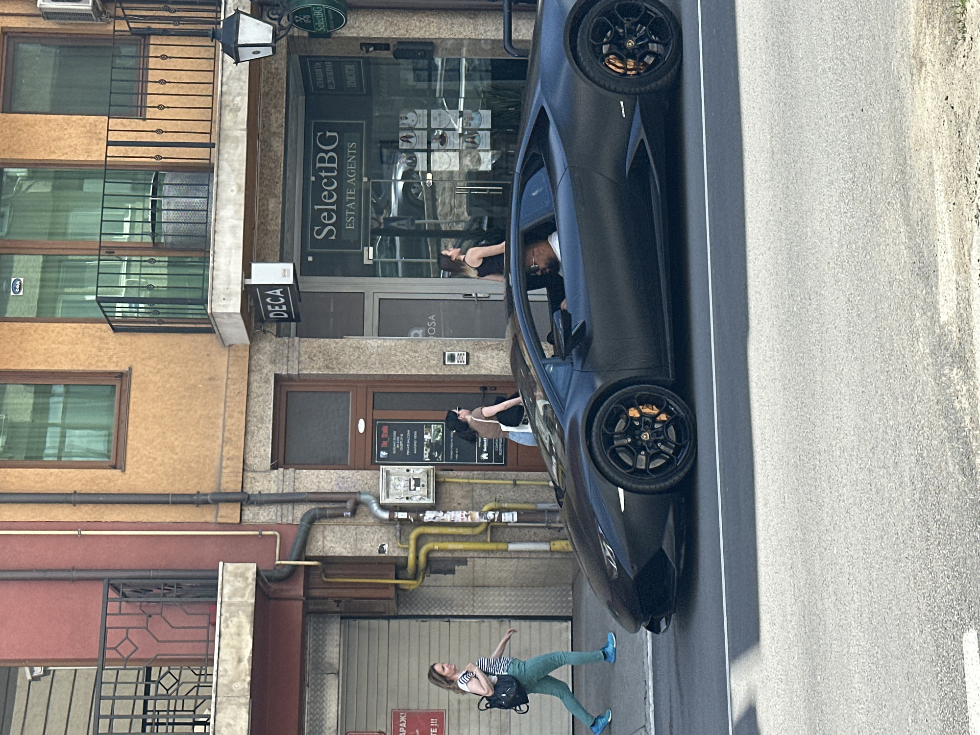 The image shows an urban street scene in Veliko Tarnovo, Bulgaria, with a black Lamborghini parked beside a sidewalk. A woman in a striped shirt and blue pants is walking away from the car. Another woman stands in the doorway of an estate agency, "SelectBG," possibly interacting with the driver, a man visible inside the vehicle. The building facades are diverse, with visible windows, balconies, pipes, and signs ("DECA," "SelectBG"). The environment also contains a grey steel roller shutter door. The street surface is paved.