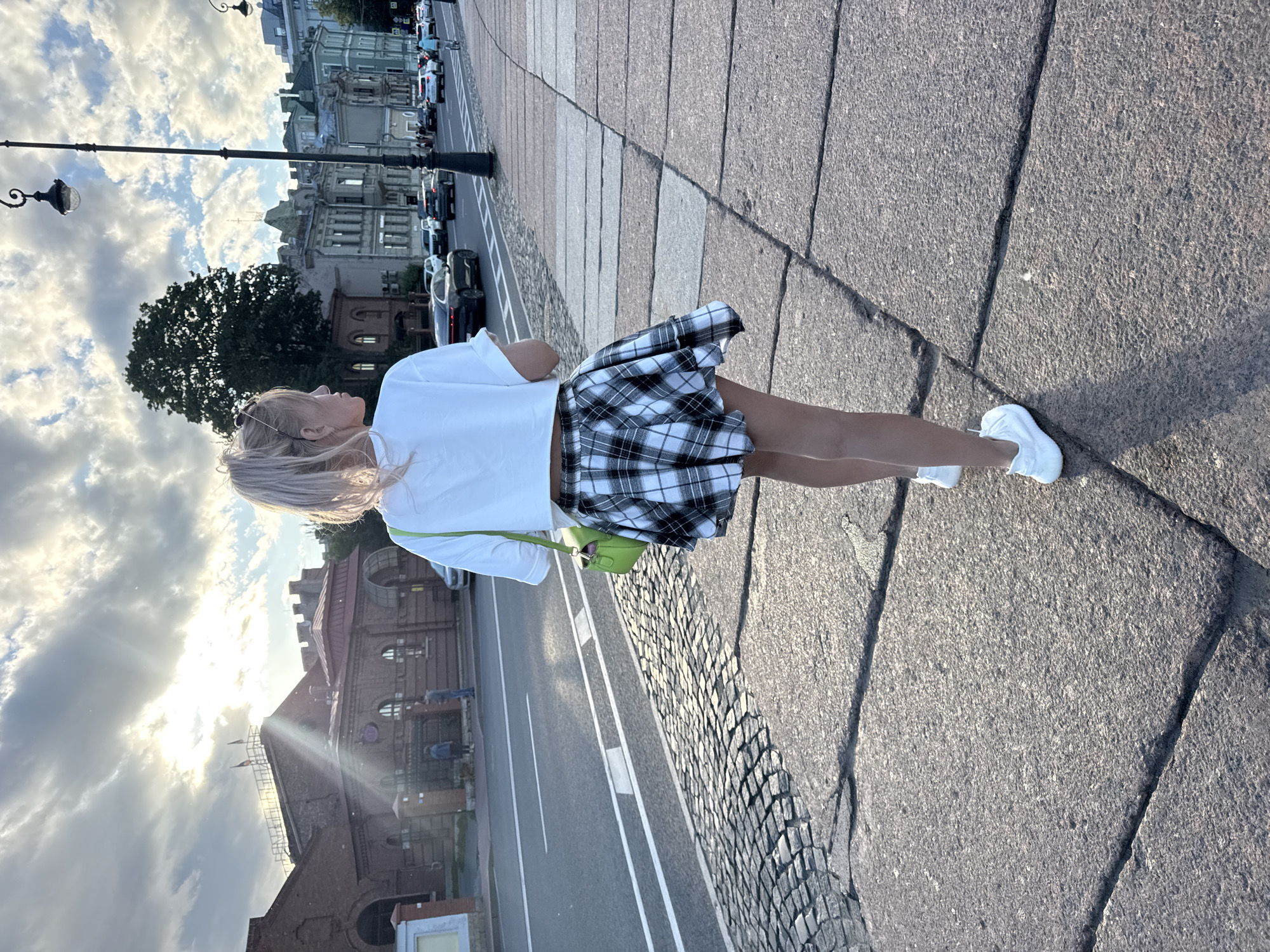 A young woman with blonde hair pulled into a ponytail walks away from the camera down a sidewalk in Saint Petersburg, Russia. She is wearing a white crop top, a black and white plaid skirt, and white sneakers. She is carrying a bright green purse on her left shoulder. The sidewalk is made of large stone slabs and is next to a street with cars driving on it. In the background, there are brick buildings with arched windows and a tree. The sky is cloudy with the sun shining through, creating a lens flare effect. This scene is consistent with a location along the Neva River embankment in Saint Petersburg.