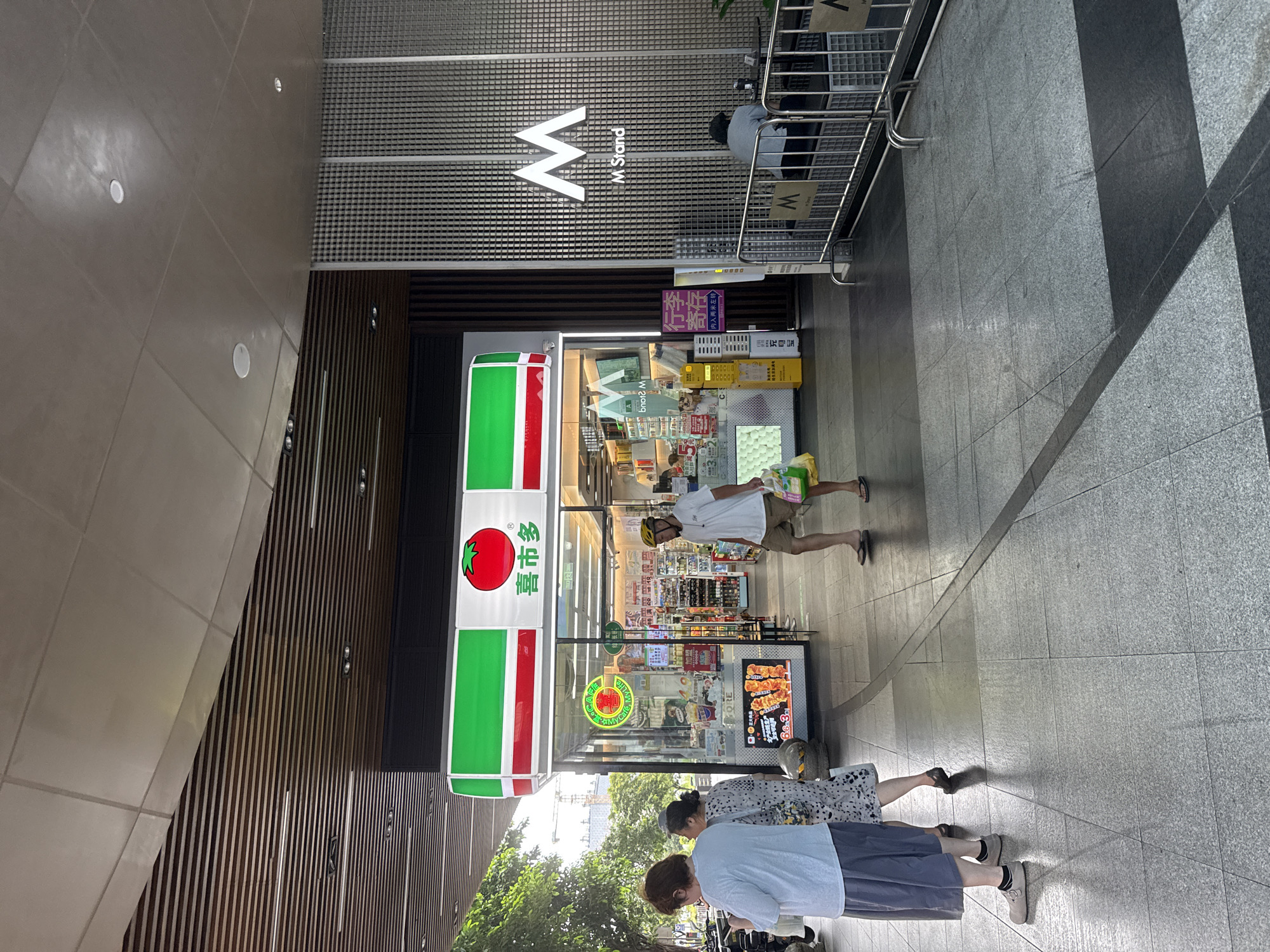In Haizhu District, China, a street-level view captures a commercial area with a 7-Eleven convenience store as the focal point. The store features a prominent red and green logo and is stocked with various items visible through its glass storefront. To the right of the 7-Eleven, an "M Stand" is advertised with a large white "M" logo on a grey patterned wall.
Several people are present in the scene. Two women are walking in the foreground, one wearing a white t-shirt and grey skirt, the other in a patterned dress. A man in a white t-shirt, khaki shorts, and a yellow helmet is walking towards the right, carrying shopping bags. Further to the right, a person is seated on a bench outside the "M Stand" area.
The environment is a paved walkway with a grey, tiled surface. The architecture includes a modern, slatted ceiling structure extending over the walkway. Shadows from the structures and overhead elements are cast on the ground. Trees and some construction activity are visible in the background. The overall atmosphere suggests a daytime scene in a bustling urban environment.