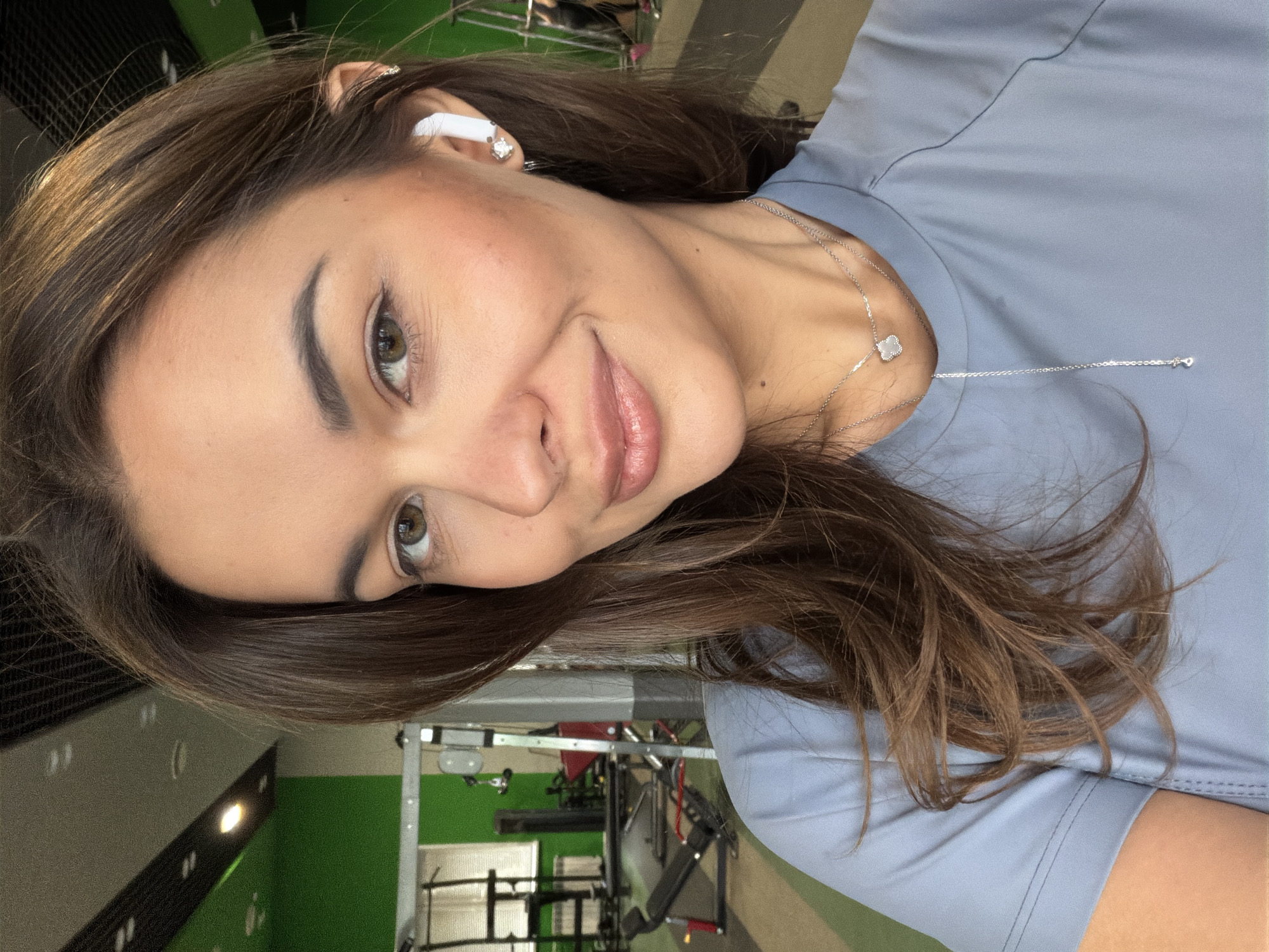 A woman with brown hair and light skin, wearing a light blue shirt and a white wireless earbud in her left ear, poses for a selfie indoors at what appears to be a gymnasium. A silver necklace with a clover pendant is visible on her neck. Exercise equipment is visible in the background. The location is potentially Krasnoyarsk, Russia, but there's no evidence to confirm this in the image.