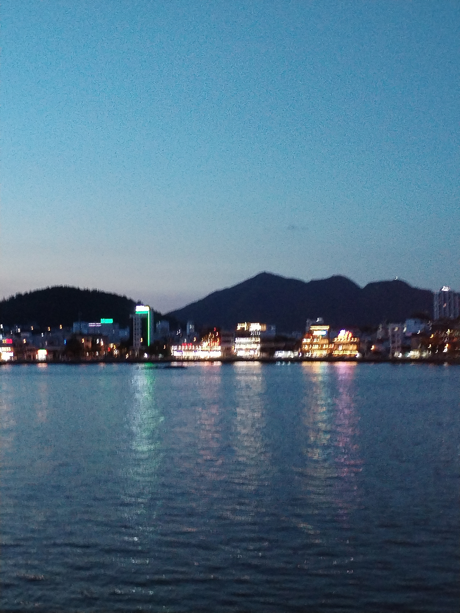 The image shows a body of water in Nha Trang Ward, Vietnam at dusk. In the background is a dark ridgeline of hills or mountains. In front of the hills, there is a line of buildings illuminated with yellow, red, and green lights that reflect in the water. The water is a dark blue-green with slight ripples. The sky is a muted, clear blue transitioning to a darker blue near the horizon. No people are visible.