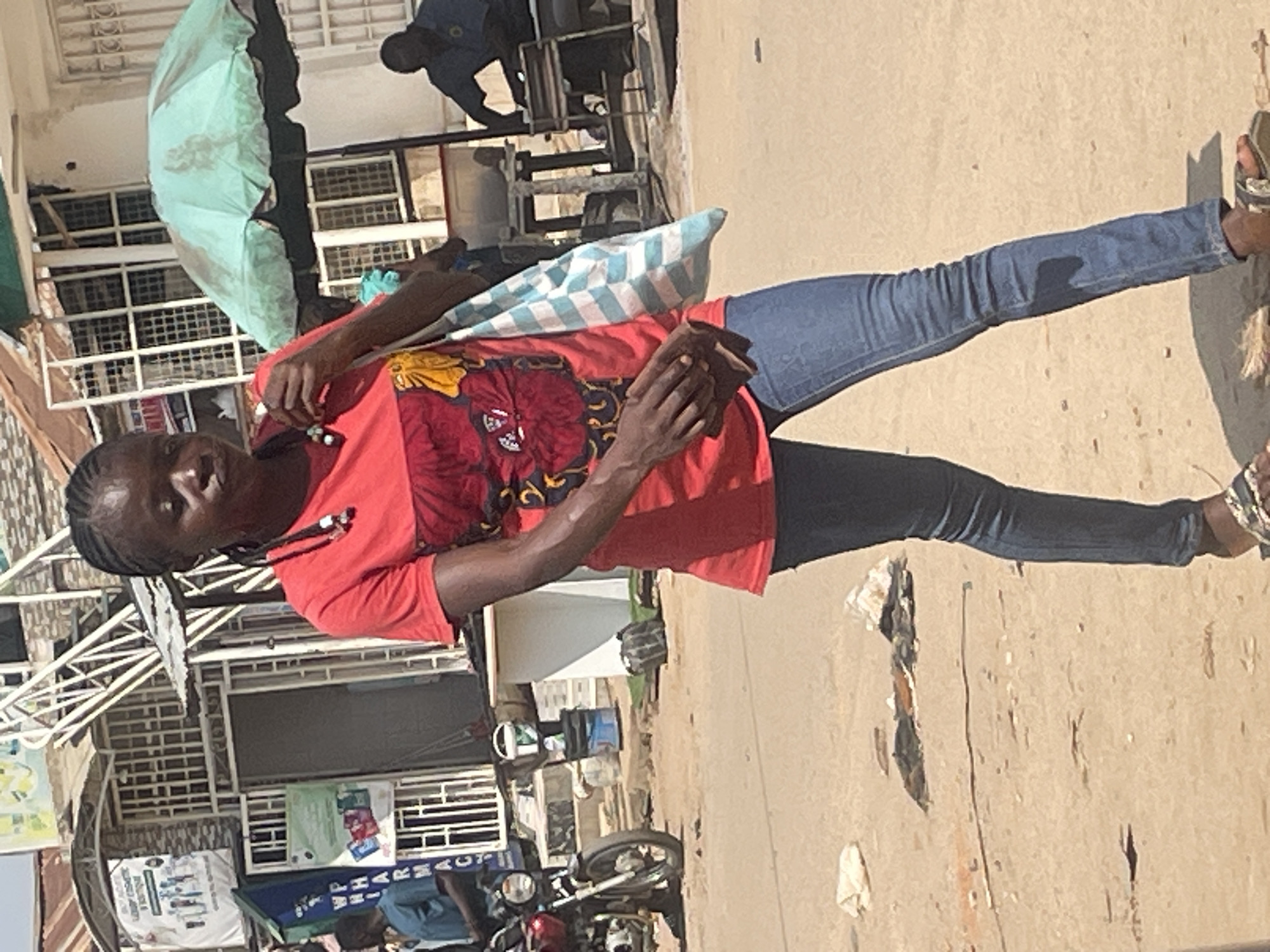 A Black woman with braided hair, wearing a red t-shirt with a floral pattern and blue jeans, walks on a dirt road in Abuja, Nigeria. She is carrying a blue and white checkered bag over her shoulder. In the background, a motorcycle is parked on the left, and a green umbrella shades a market stall on the right. Several buildings and people are visible in the distance, suggesting a busy street scene.
