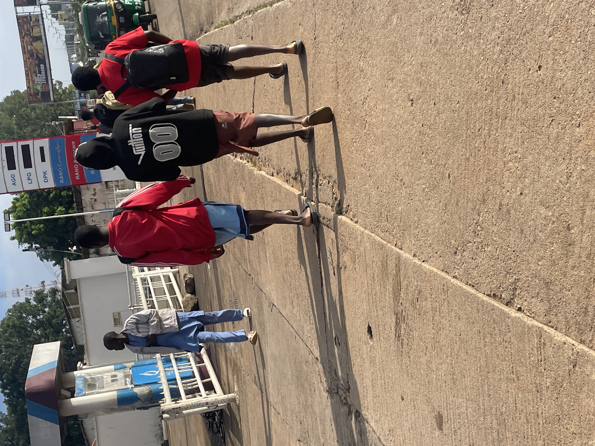 The image shows a sunny day at a gas station in Jos, Nigeria. In the foreground, three young men are walking away from the camera on a concrete sidewalk. The man on the left is wearing a red jacket and blue shorts, and carries a backpack. The man in the middle is wearing a black hoodie with "Vantion 00" written on the back, and brown shorts. The man on the right is wearing a red t-shirt and black shorts, and also carries a backpack. Behind them, a gas station with several pumps is visible, as well as a white fence and a building. In the background, trees and a transmission tower can be seen. A green three-wheeled vehicle is also visible on the road. The overall scene depicts a busy urban environment on a clear day.