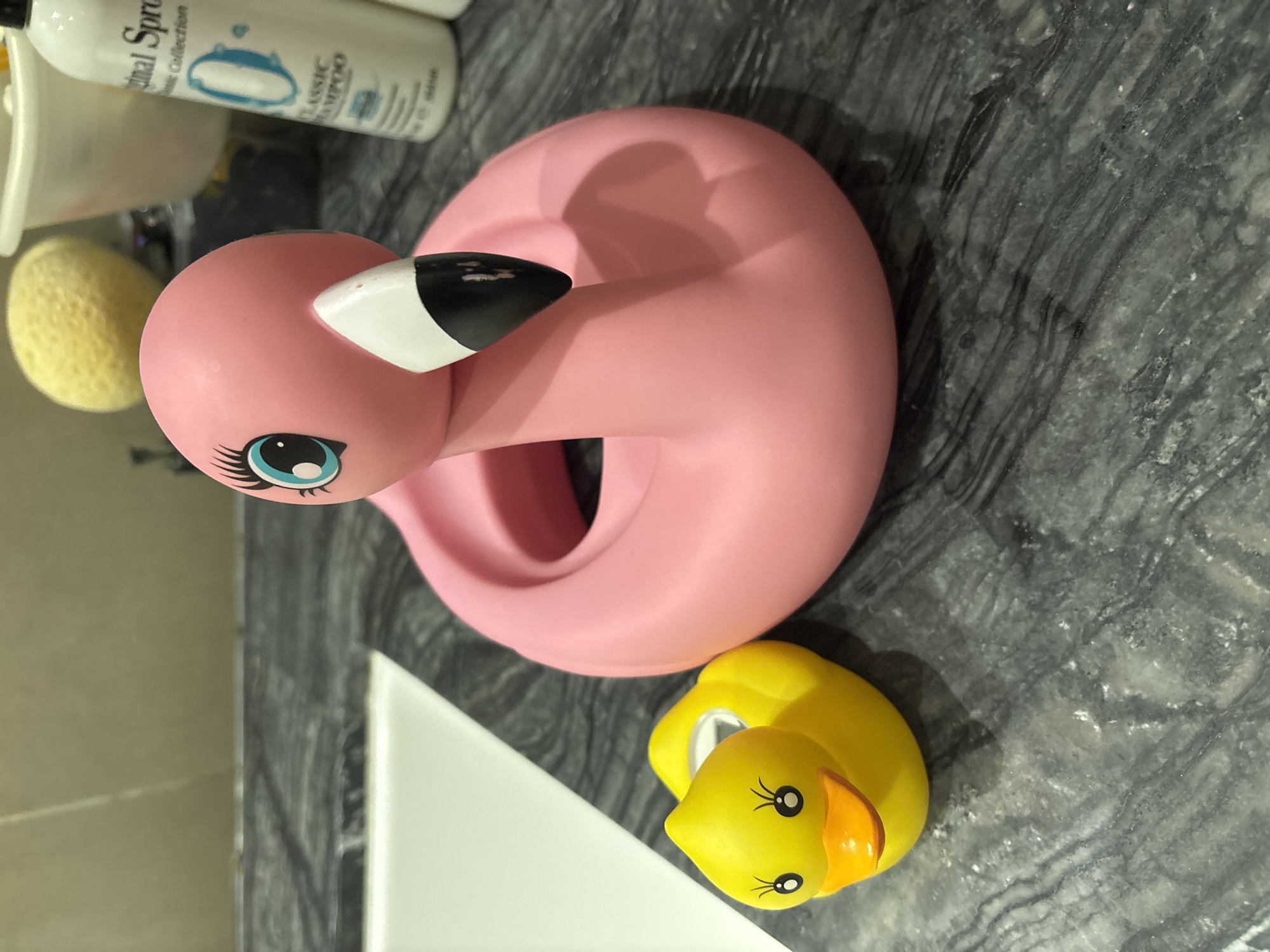 This is a close-up, eye-level shot of two rubber bath toys, a yellow duck and a pink flamingo, resting on a dark gray, marbled countertop. The duck is to the left of the flamingo. The flamingo is larger, its body forming a pink ring with a neck and head that extends upwards, featuring large blue eyes and a black and white beak. Behind the toys, a white sink is partially visible on the left, and on the right, a bottle of "Original Sprout Classic Shampoo" and a yellow loofah are present. The overall scene suggests a bathroom or vanity area. There is no indication of Dubai, United Arab Emirates.