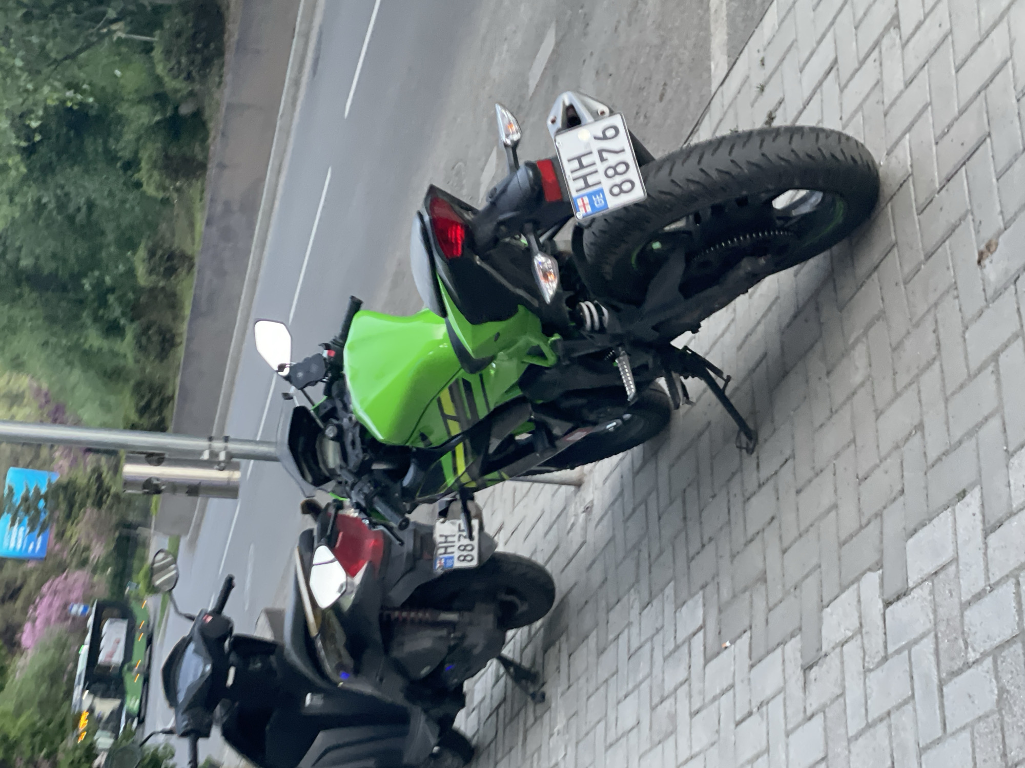 The image depicts two motorcycles parked on a paved sidewalk in what appears to be a public space. One motorcycle is a newer, green Kawasaki Ninja, while the other is a black scooter. The license plate of the Kawasaki Ninja reads "HH 8876". The surrounding environment includes a road, some trees and greenery, and a street light. Based on the license plate and general appearance of the area, it's possible that the photo was taken in a European city, potentially Tbilisi, Georgia.