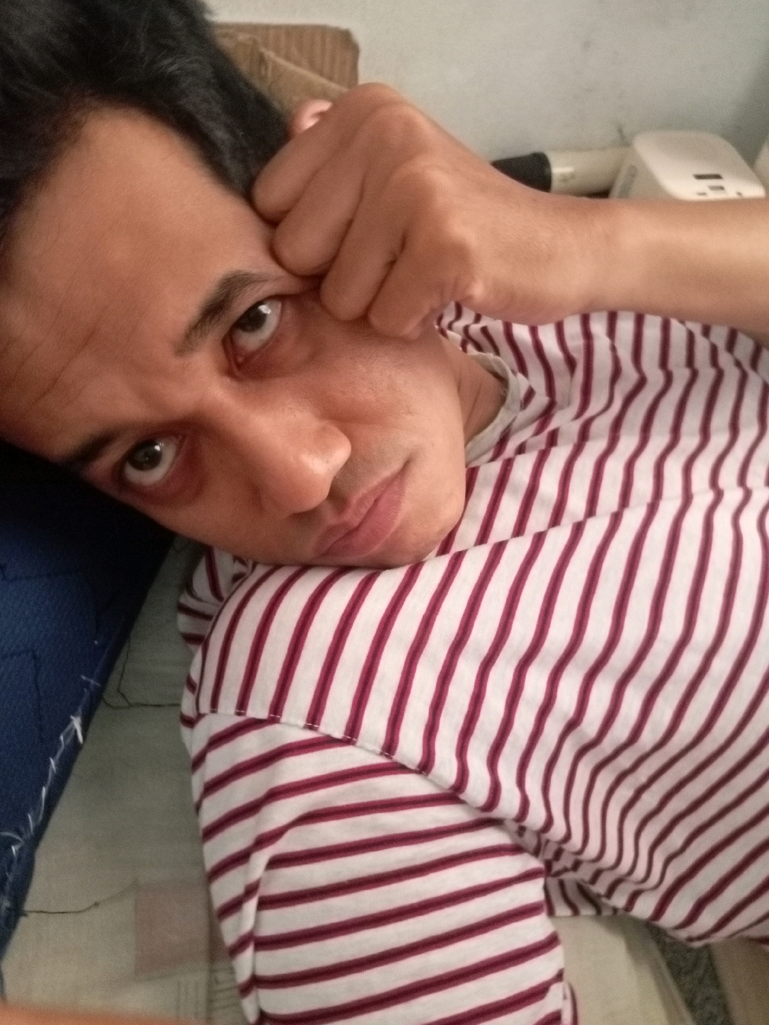 The image shows a close-up, head-and-shoulders view of a person lying down, resting their forehead and eye with their hand. They are wearing a striped shirt with alternating maroon and white stripes. The background is blurred but suggests an indoor setting, possibly a bedroom or living area, with a plain wall and some objects on a shelf. The person's gaze is directed towards the camera. The lighting is soft, creating a relaxed and intimate atmosphere. No specific location in Batam, Indonesia is discernible from the image itself.