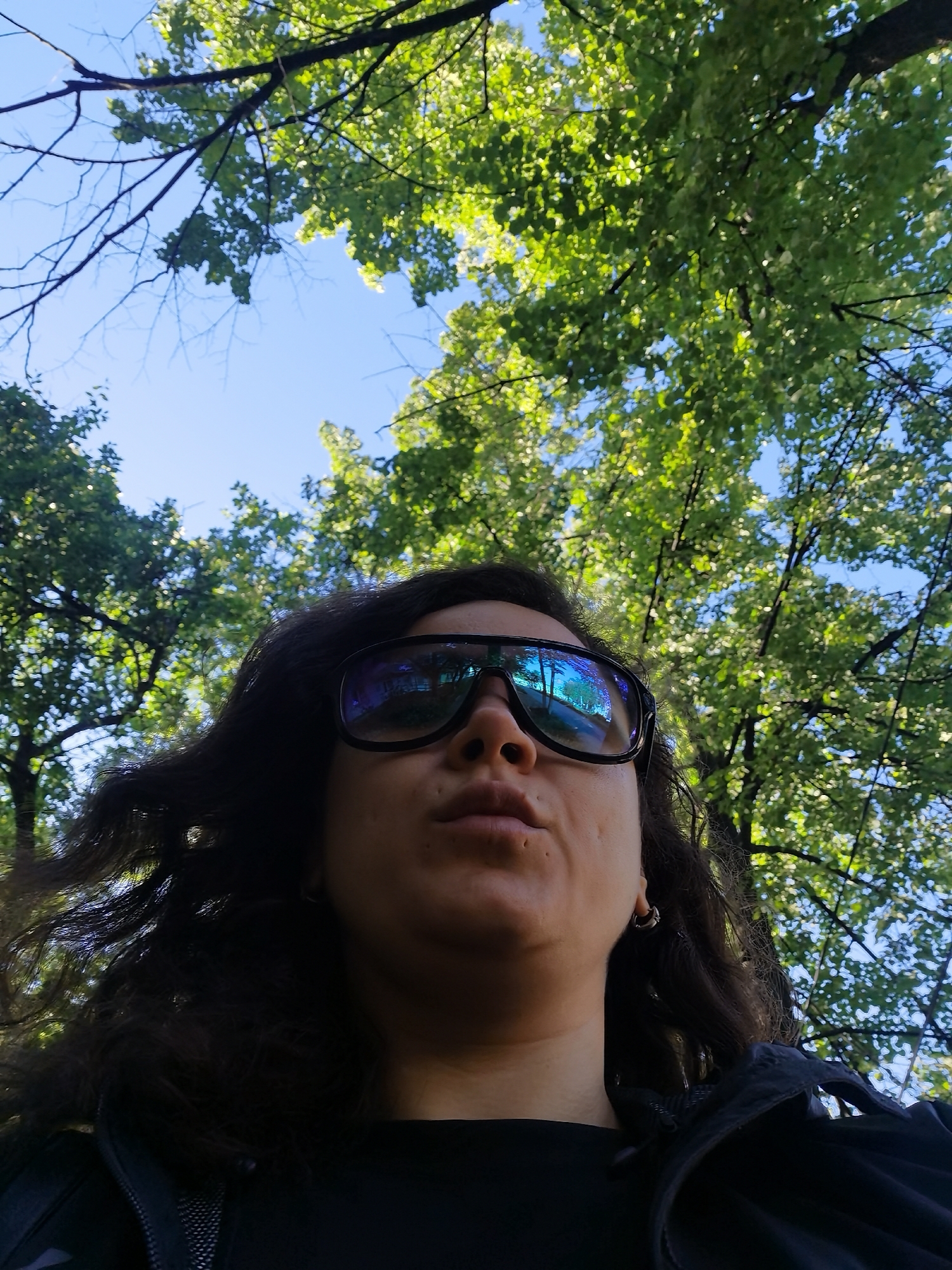 A low-angle, medium shot captures a person looking up through a canopy of green trees on a bright, sunny day. The person, wearing large, dark sunglasses with blue reflective lenses, has dark, curly hair and appears to be wearing a black top and jacket. The sky is clear and blue, with sunlight filtering through the leaves, creating a dappled effect. The image provides no specific information to determine the location in Chaikovsky, Russia.