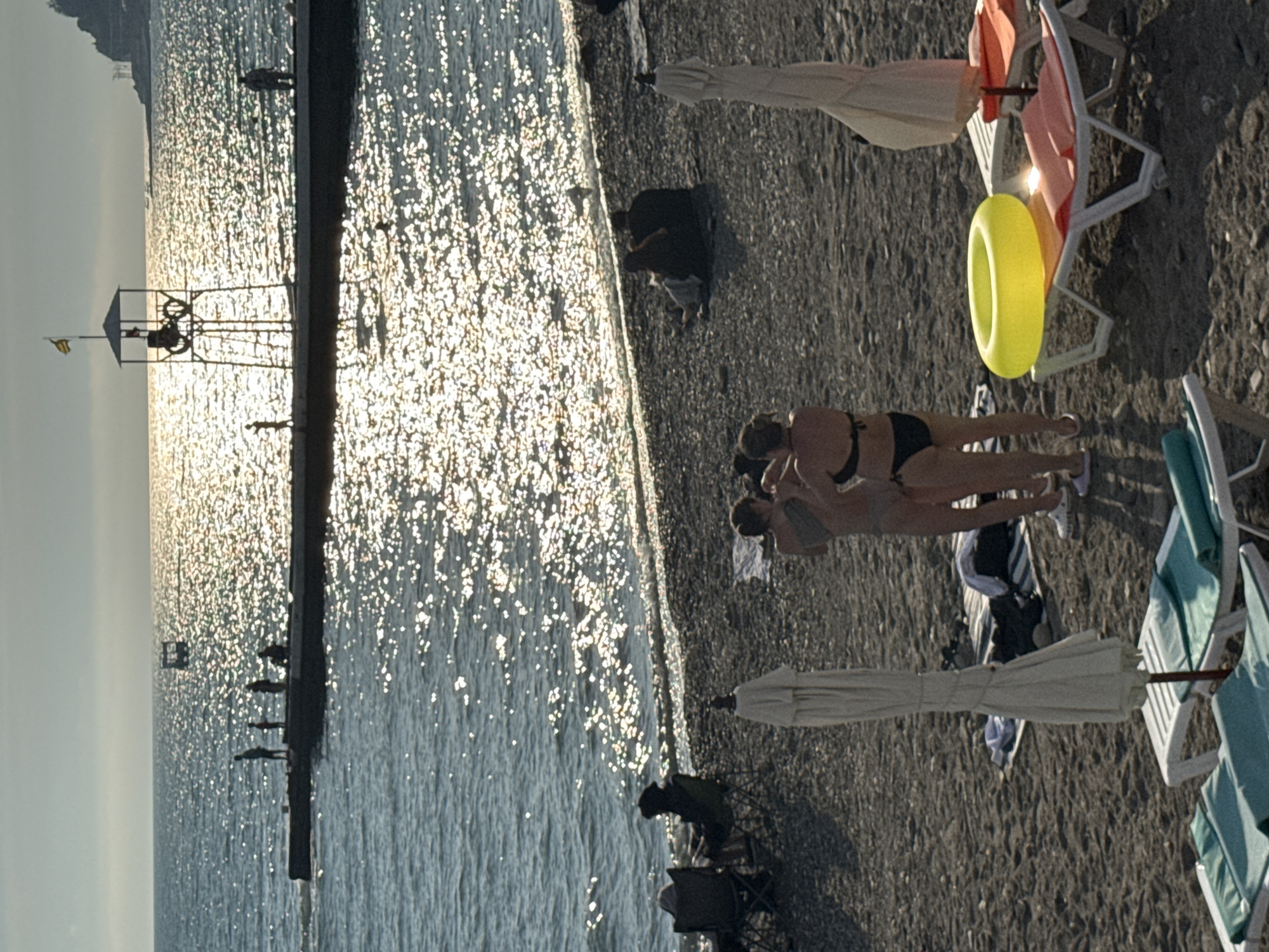 The image depicts a beach scene in Sochi, Russia, with a view of the Black Sea under a bright, sunny sky. The foreground shows a section of a pebbly beach with several beach chairs, some with towels and belongings, and two closed beach umbrellas. In the middle ground, two individuals, a woman in a bikini and another person, are standing near the water's edge, facing away from the camera. Further out on the water, a long pier extends into the sea. Several silhouetted figures are visible on the pier, some standing and some seated. In the background, a lifeguard tower stands on the pier, and a small boat can be seen in the distance. The sun's reflection creates a shimmering effect on the surface of the water.