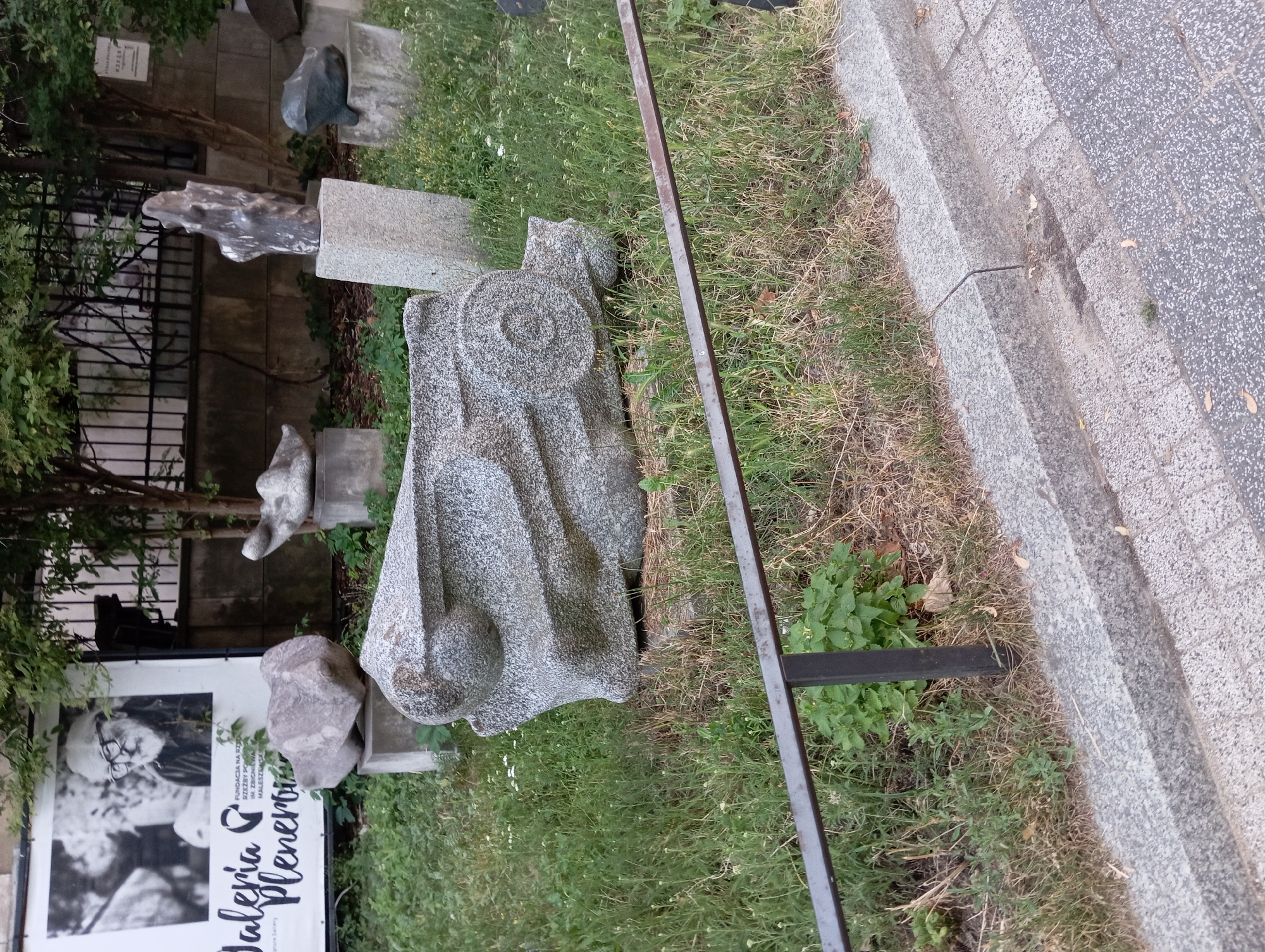 The image depicts an outdoor art installation featuring several abstract sculptures made of stone. The sculptures are situated in a grassy area with some dry patches and scattered white flowers, bordered by a grey granite curb and a cobblestone sidewalk. In the background, a black metal fence separates the art area from a wall. A large banner with a black and white portrait of an elderly man with glasses and text in Polish is visible on the left. The overall scene suggests an exhibition or public display of art in an urban environment. The sculpture in the foreground is large, grey, and textured, with a design that resembles mechanical parts or an abstract form. Other sculptures in the background are smaller and placed on pedestals. The environment is an outdoor public space, likely in a park or gallery setting in Warsaw, Poland, as indicated by the Polish text on the banner.