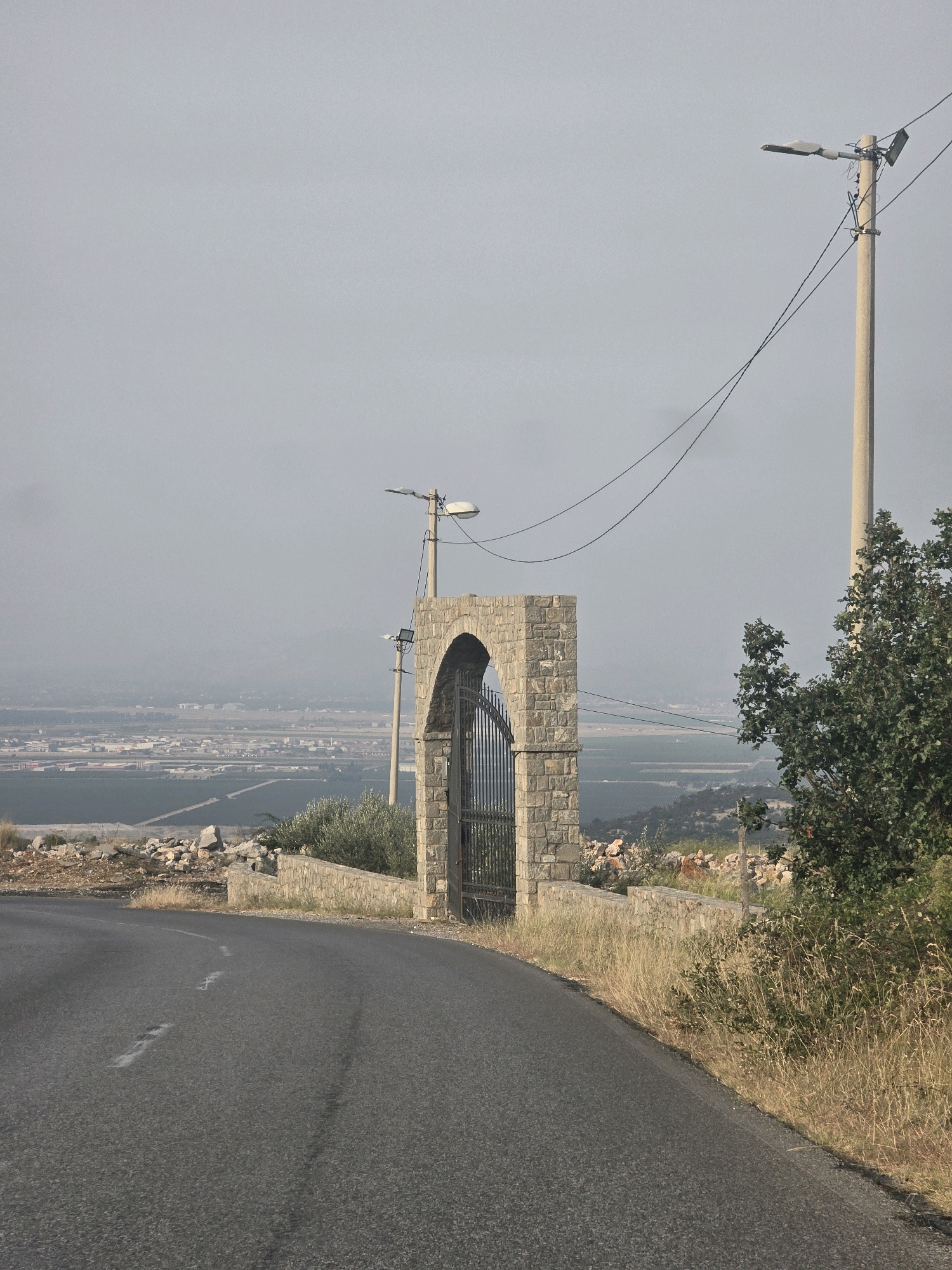 A paved road curves to the right in the foreground, with dry grass and rocks on the right side of the road. A stone archway with a metal gate stands on the right side of the road. Beyond the archway, a distant landscape with buildings and fields is visible under a hazy sky. A utility pole with a light fixture stands to the right of the archway, and another pole with a light fixture is visible between the archway and the distant landscape. A tree with green leaves is situated to the right of the archway.