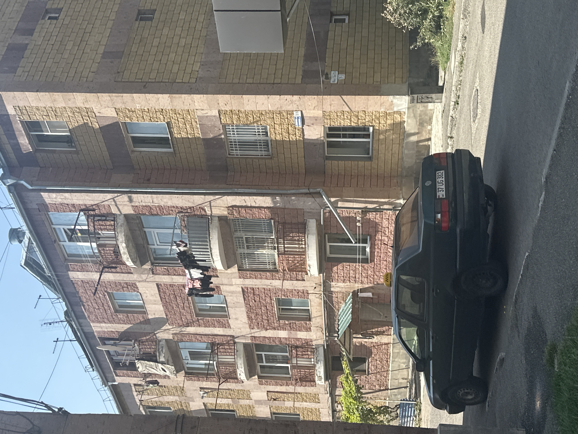 The image shows a dark green Volkswagen sedan with a taxi sign on its roof, parked on a street in Gyumri, Armenia. The car's license plate reads "37 UG 382". To the right of the car is a multi-story apartment building with a brick facade, featuring balconies with laundry hanging out to dry. The building's design incorporates alternating bands of red and light yellow brick. The street appears to be asphalt, with a sidewalk and some greenery visible. The sky is clear and blue, indicating a sunny day. Several antennas are visible on the rooftop of the apartment building. Street signs are also visible on the building's facade.