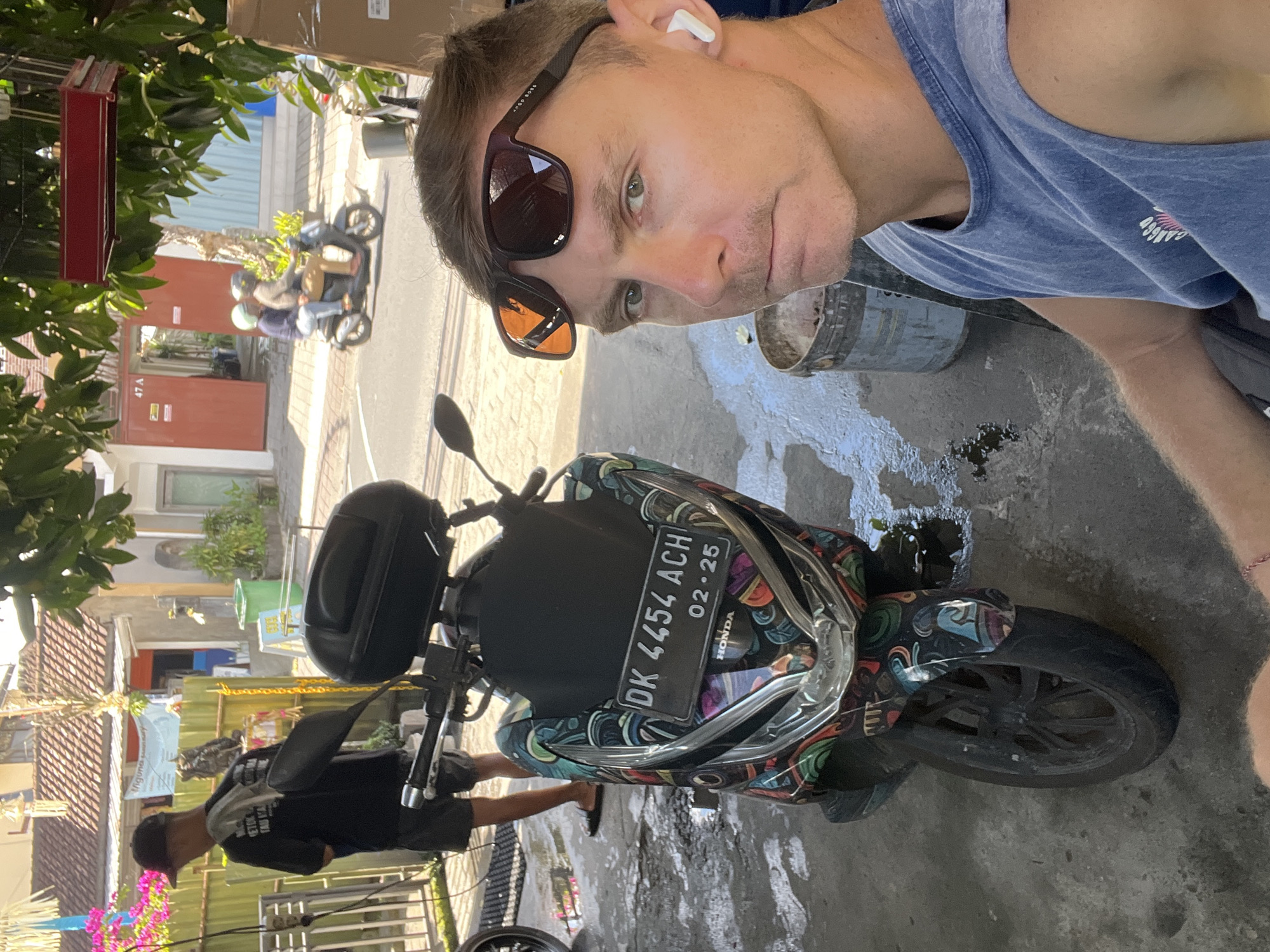 A man with a tan complexion and short brown hair is taking a selfie outdoors in a city environment, possibly North Kuta, Indonesia. He is wearing a blue tank top and sunglasses, with a white earbud in his right ear. To his left, a colorful, decorated scooter is parked on a wet concrete surface. In the background, there is a person wearing a black t-shirt and shorts, standing near a fence. Further in the background, buildings and two people riding a scooter are visible. The overall scene appears to be a street or alleyway with some greenery and a sense of everyday life.